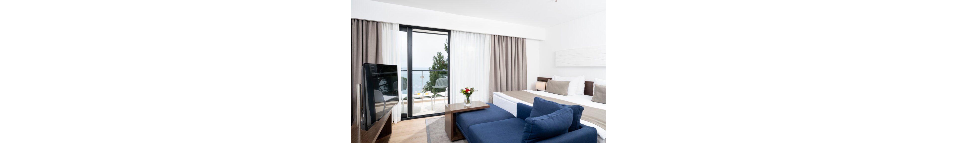 Bright contemporary hotel room featuring a double bed and blue sofa. Large floor-to-ceiling windows open to a balcony with outdoor seating and a view of the sea and trees. Neutral white and beige walls and curtains create a clean, minimalist atmosphere. A television, wooden furniture, and a vase of flowers add comfort and decorative accents.