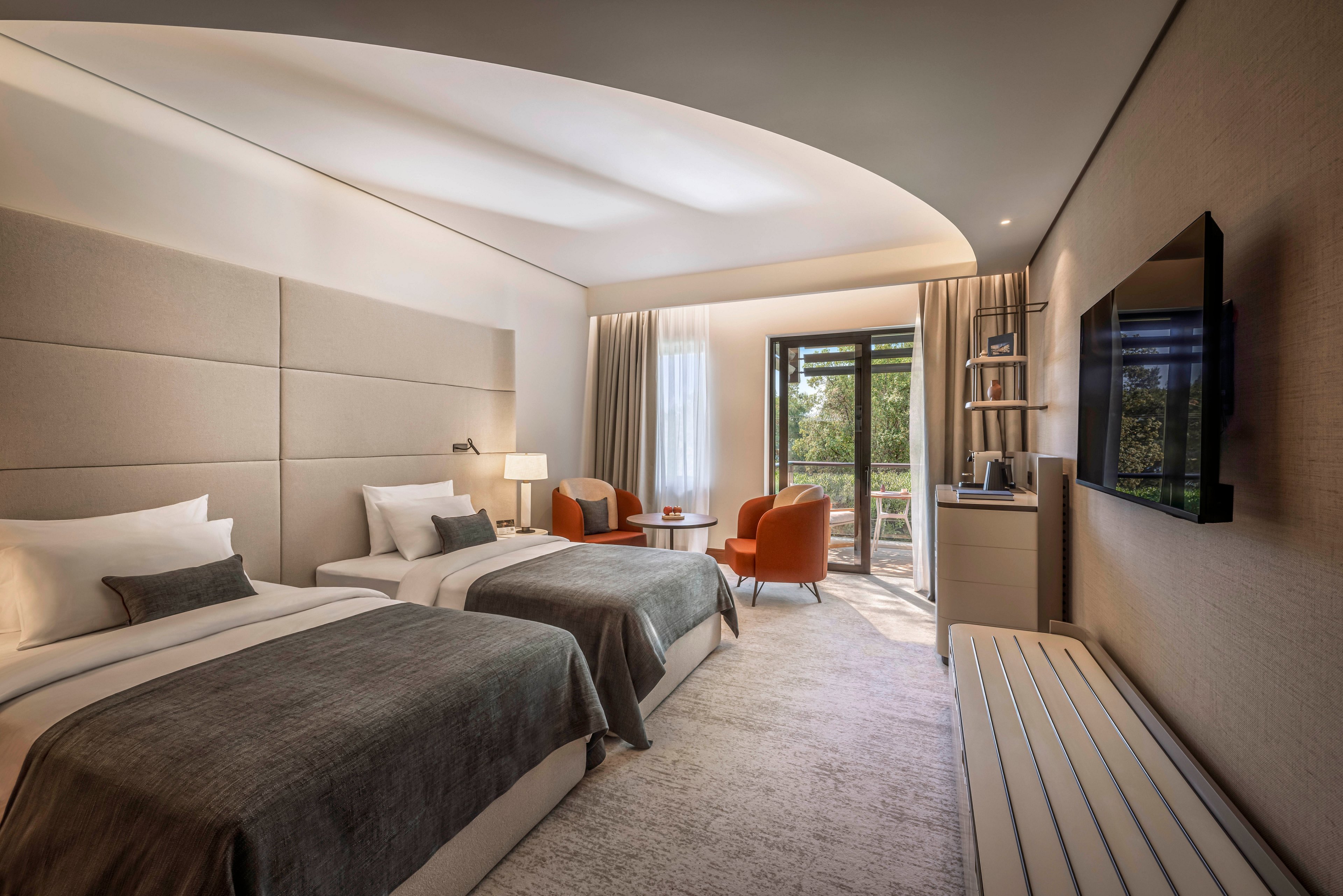 Contemporary hotel room featuring two neatly made twin beds with gray throws and white linens. A seating area with two orange armchairs and a small table is positioned near the window and balcony door. Soft, indirect ceiling lighting and neutral wall tones create a calm, upscale atmosphere. A flat-screen television, desk, and minibar complete the modern guestroom amenities, while natural light filters in from the balcony overlooking greenery.