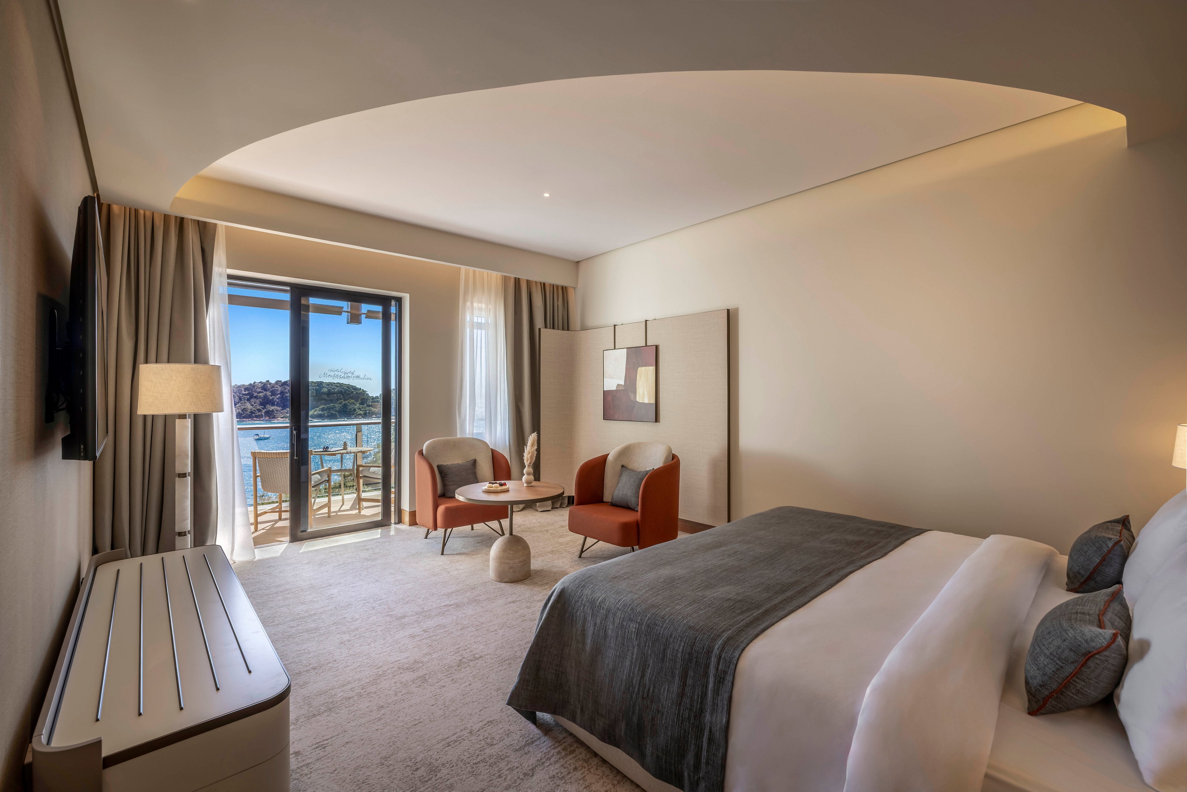 A contemporary hotel bedroom features a large neatly made bed with gray and white linens. The room includes a seating area with two armchairs and a small table near floor-to-ceiling glass doors. Outside, a balcony overlooks a scenic waterfront and distant hills under clear daylight. Neutral walls, soft lighting, and minimalist decor create a calm, upscale atmosphere.