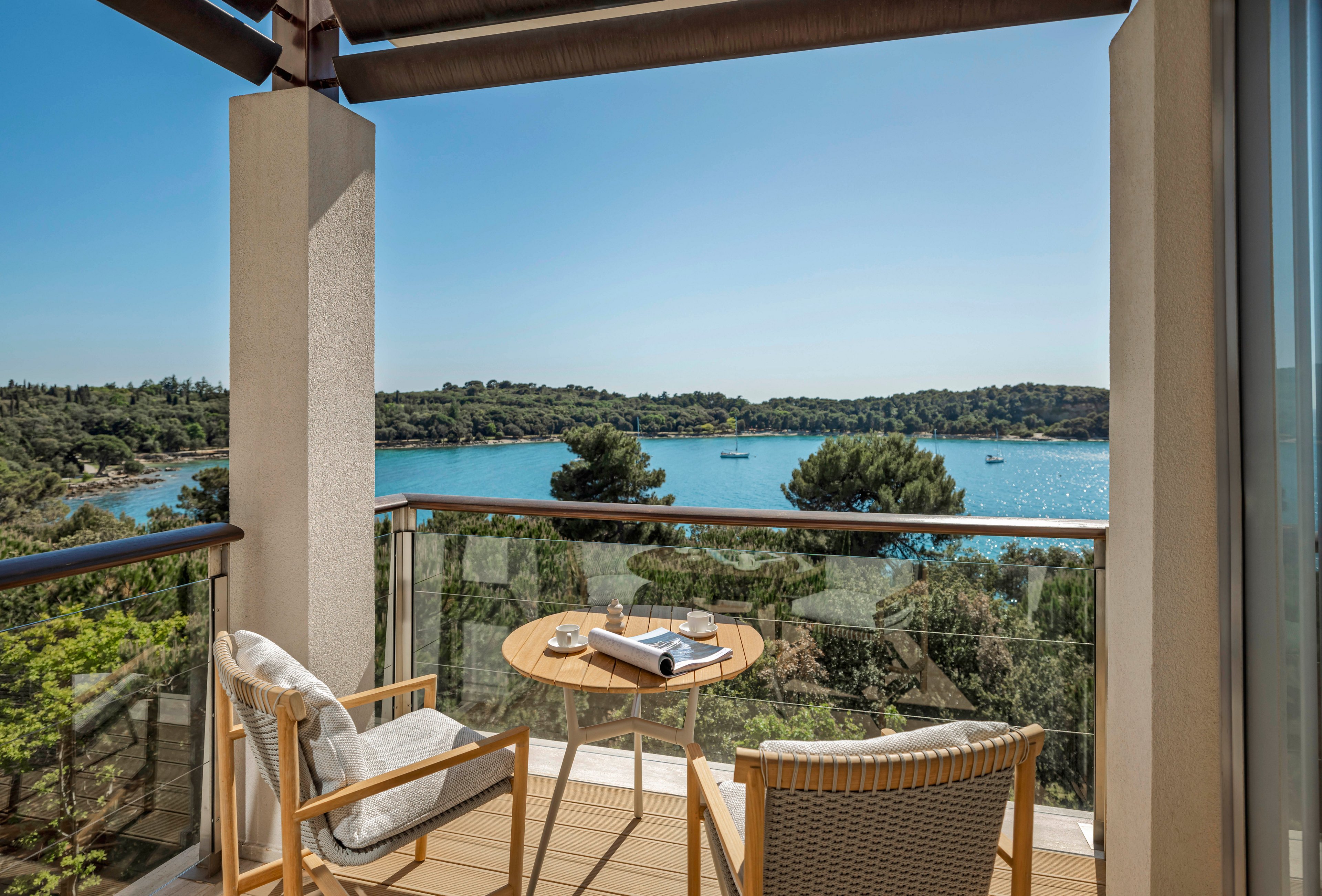 A modern balcony terrace features two wooden chairs and a small round table set for relaxation. The space opens onto a stunning view of a calm blue bay dotted with anchored boats. Lush green trees and a distant shoreline frame the water under a clear, cloudless sky. The scene suggests a peaceful vacation setting ideal for reading, resting, or enjoying the coastal landscape.