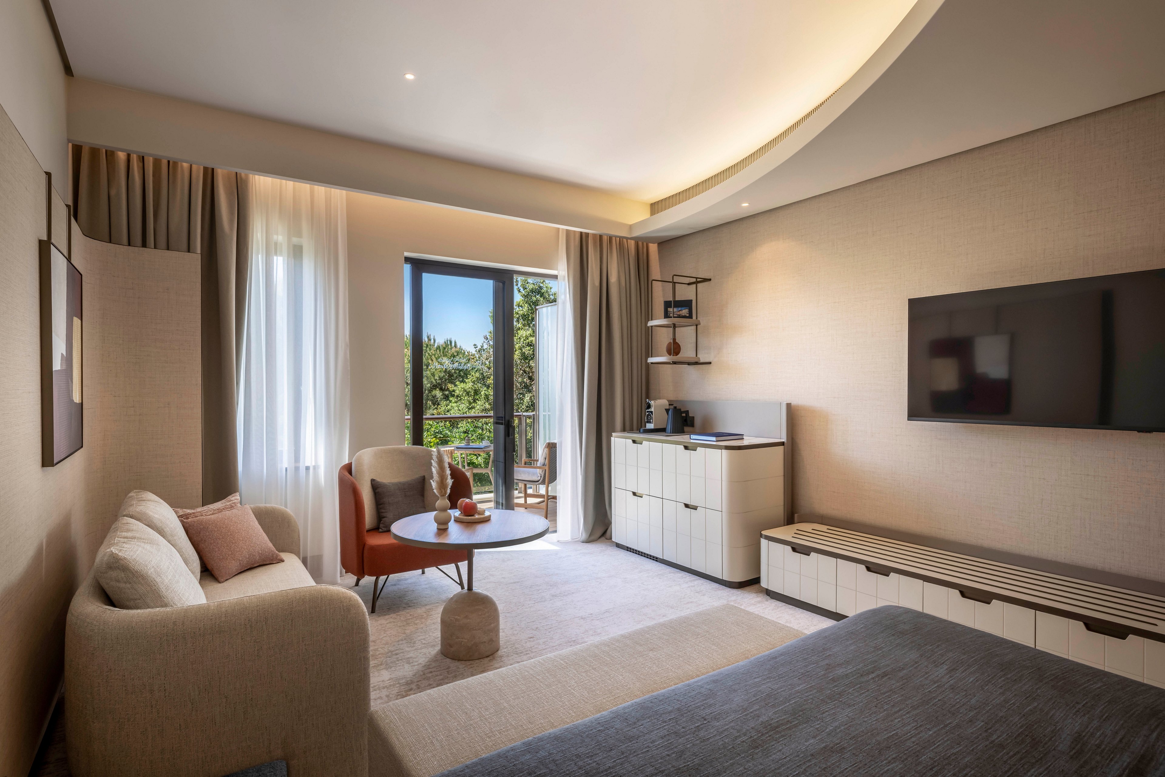 Contemporary hotel room interior featuring a cozy sofa, round table, and large bed. A wall-mounted flat-screen TV faces the seating area, with built-in cabinets and open shelving along one wall. Floor-to-ceiling curtains frame a glass door that opens to a balcony overlooking greenery. Soft neutral tones, indirect ceiling lighting, and minimalist decor create a calm, inviting atmosphere.