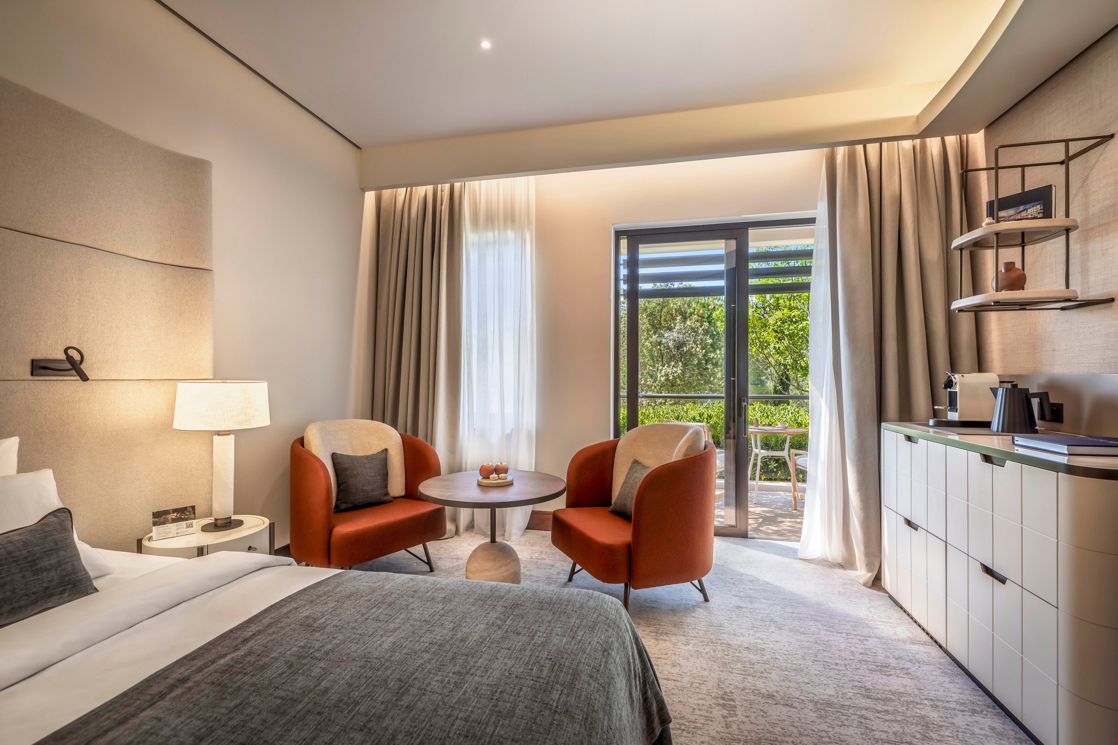 Contemporary hotel room interior featuring a neatly made bed and soft neutral tones. Two orange armchairs and a round table create a cozy sitting area near the window. Large glass doors open to a terrace with greenery outside, allowing natural light to fill the space. Built-in cabinetry, open shelving, and a coffee station add functional comfort to the modern design.