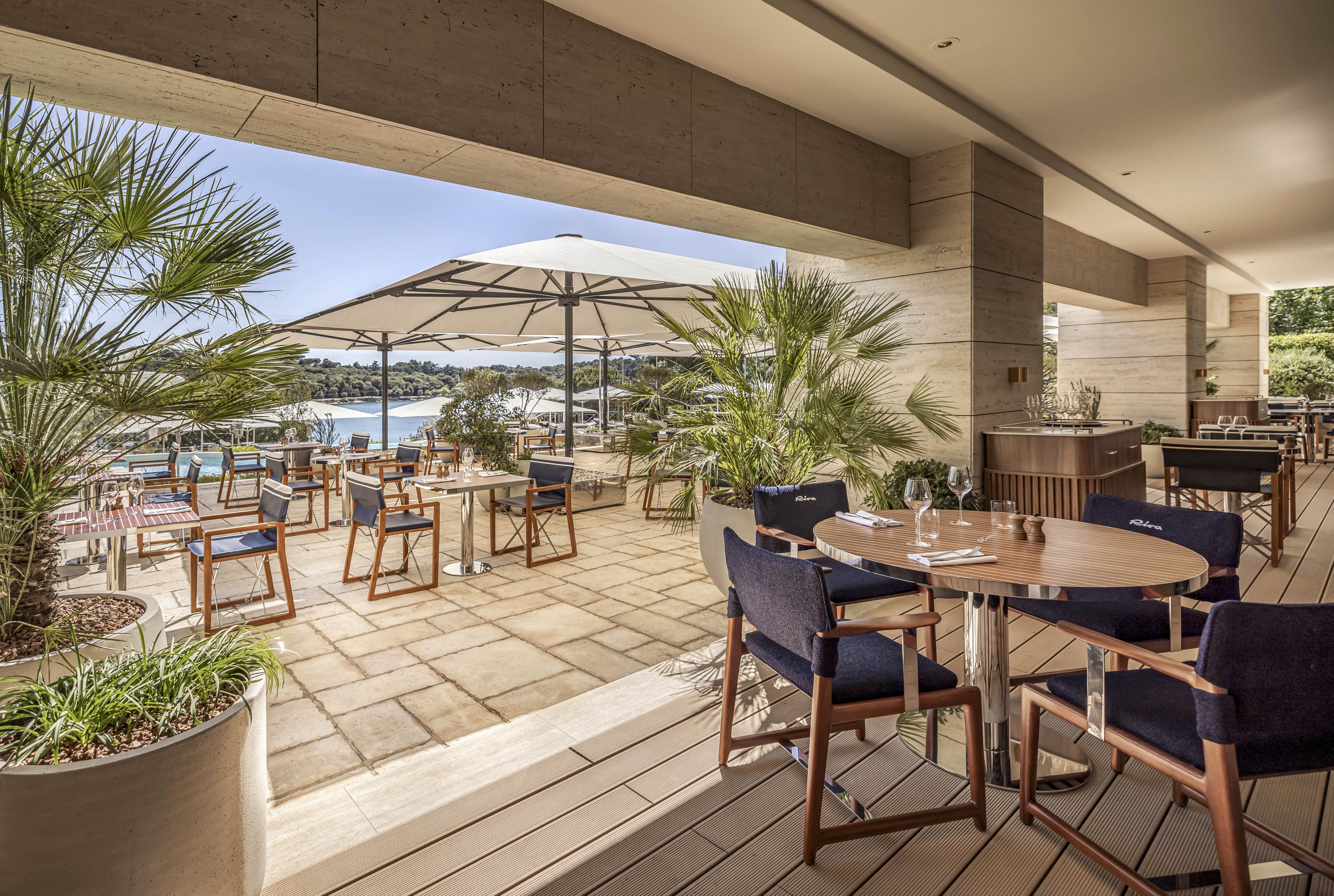 An upscale open-air restaurant terrace overlooks a scenic waterfront. Wooden tables and navy cushioned chairs are arranged neatly under large white patio umbrellas. Potted palm trees and greenery frame the stone-tiled floor, creating a relaxed Mediterranean atmosphere. Sunlight fills the space, highlighting the inviting outdoor dining setup and distant boats on the water.