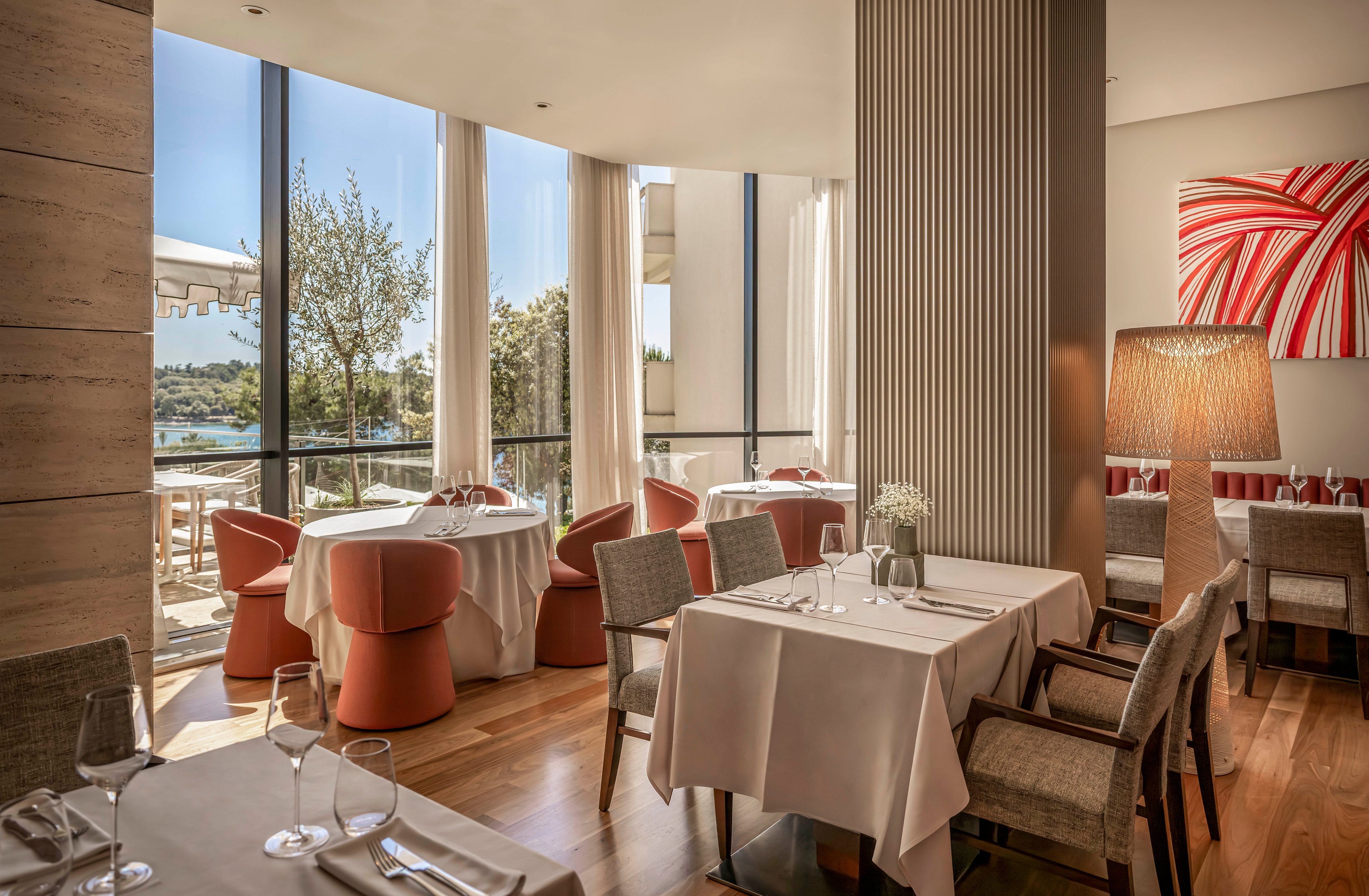 Elegant restaurant dining room with neatly set tables and upholstered chairs is bathed in warm natural light from large floor-to-ceiling windows. Round tables with coral-colored armchairs and square tables with neutral-toned chairs create a modern, comfortable seating arrangement. A tall textured column, large floor lamp, and abstract red and white wall art add contemporary design accents. Outside, a terrace with greenery and a distant water view enhances the airy, upscale atmosphere.