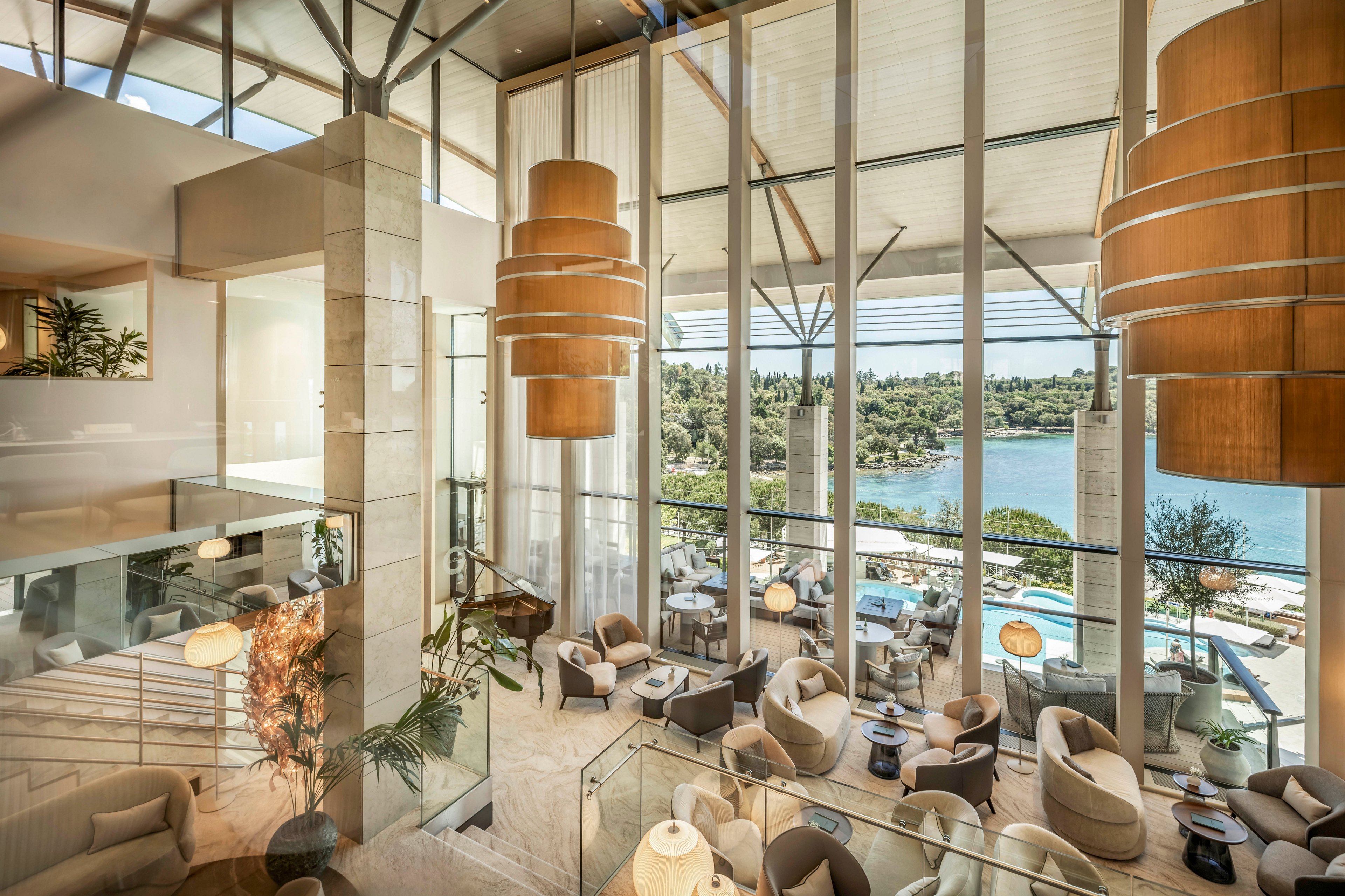 Spacious contemporary hotel lounge with large floor-to-ceiling windows and comfortable seating areas. Warm wooden pendant lights and neutral-toned armchairs create an inviting atmosphere. The interior opens to a scenic view of a blue bay, green trees, and outdoor terraces below. Natural light floods the space, highlighting the modern architecture and relaxed resort setting.