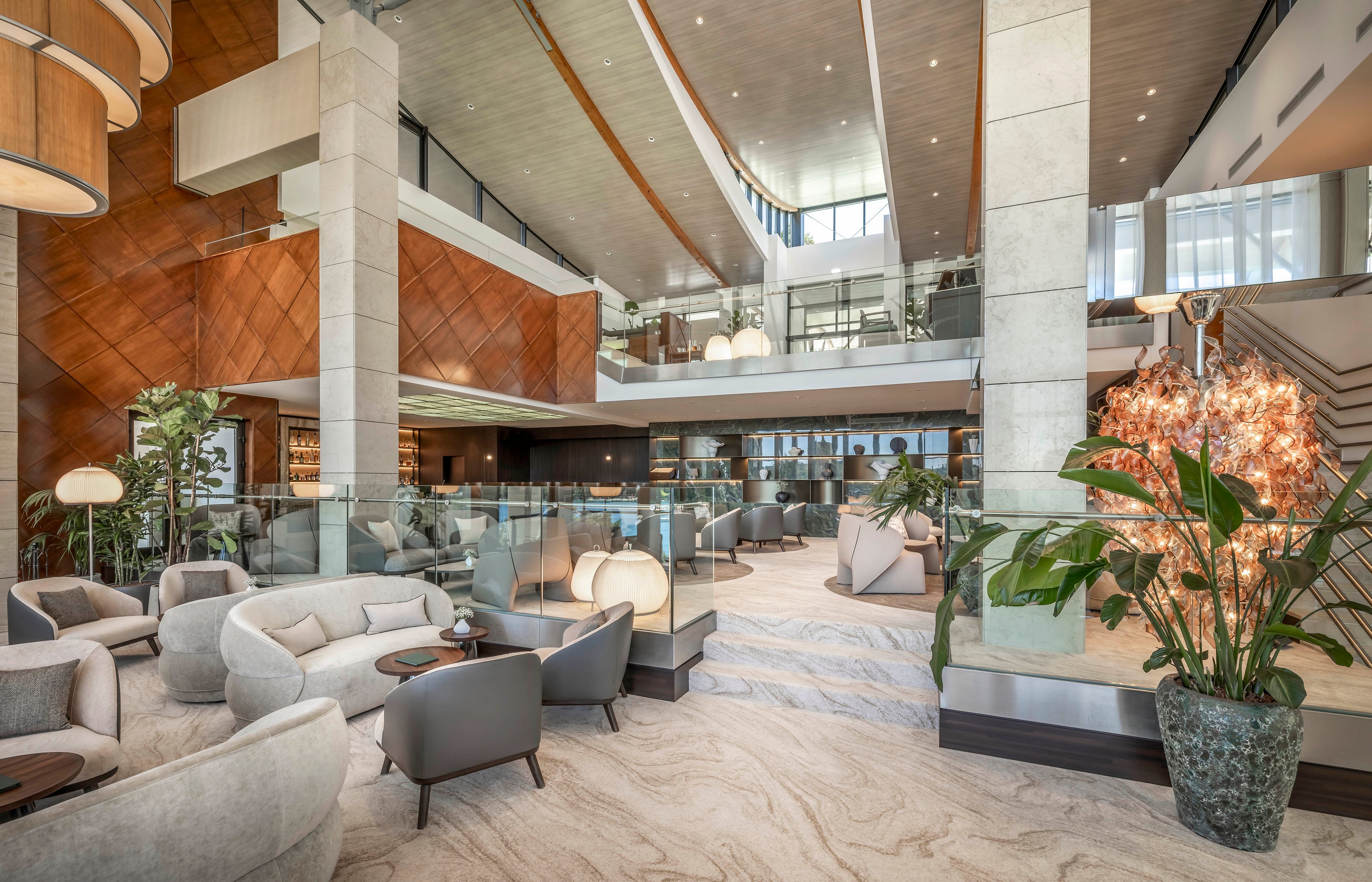 Large modern lobby lounge with plush seating and glass railings is shown in a bright, open space. Neutral-toned armchairs and sofas are arranged in conversational groupings on a light marble-patterned floor. Tall columns, wood paneling, and layered ceilings add architectural interest, while plants and decorative lighting provide warmth. A mezzanine level and floor-to-ceiling windows enhance the airy, upscale atmosphere.