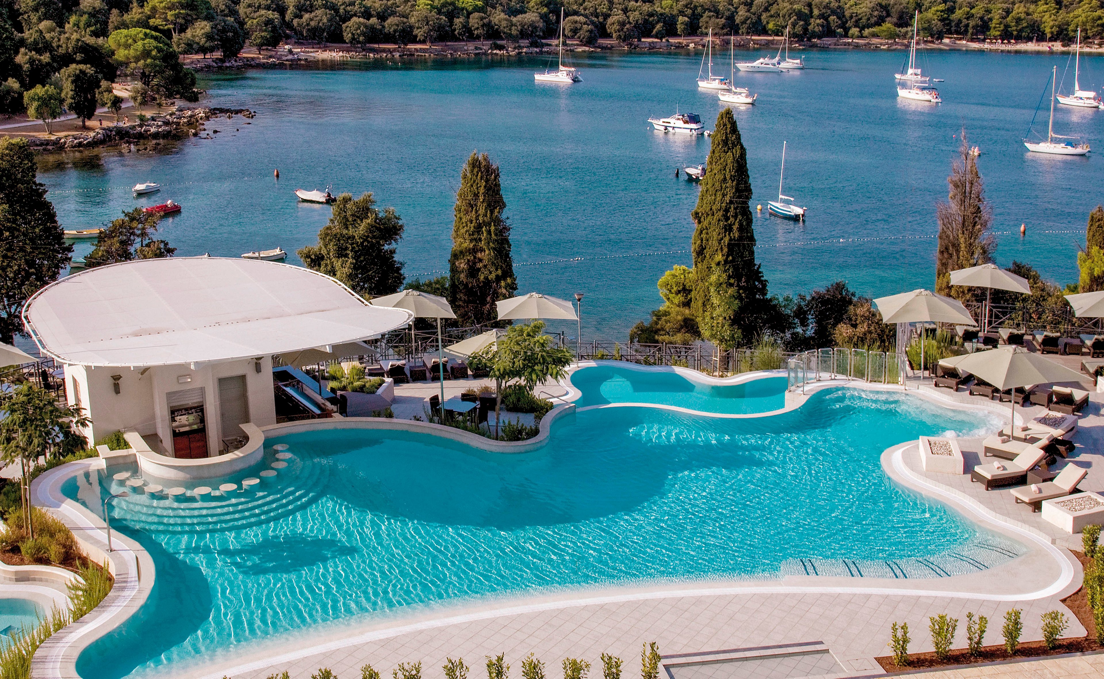 A large, curving outdoor swimming pool sits beside a calm blue bay dotted with anchored sailboats. The pool area features a swim-up bar under a white canopy and several shaded lounge areas with umbrellas and sunbeds. Lush greenery and tall trees surround the resort, creating a secluded coastal atmosphere. The scene suggests a relaxing vacation setting with clear water, modern design, and scenic ocean views.