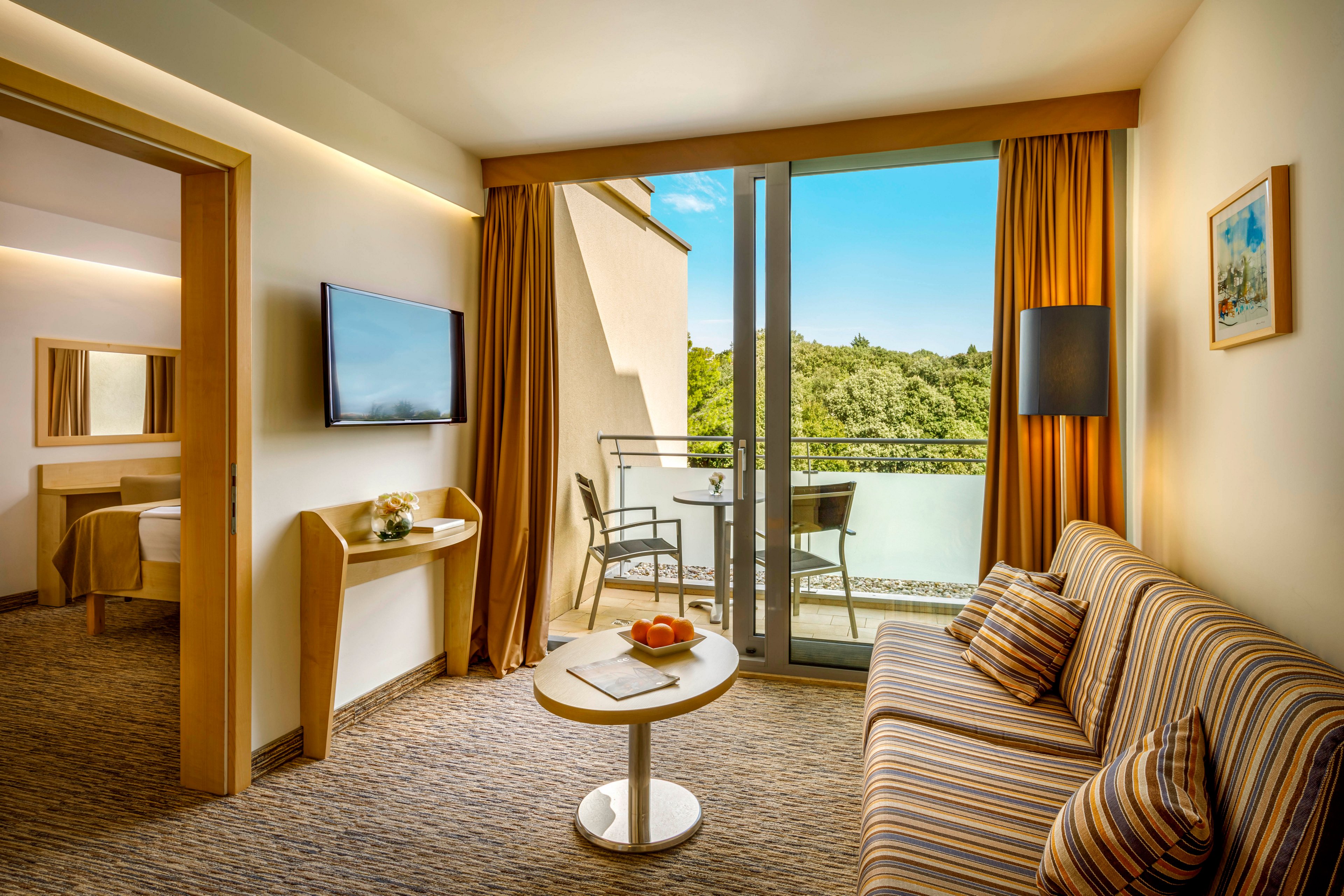 A contemporary hotel suite features a cozy sitting area with a striped sofa and a small round table holding a bowl of fruit. Warm yellow curtains frame large glass doors that open onto a balcony with two chairs and a table overlooking lush green trees and a river or lake. A wall-mounted flat-screen television and minimalist wooden furniture create a clean, modern look. In the adjoining room, a neatly made bed and a bright bathroom mirror are visible, enhancing the sense of comfort and relaxation.