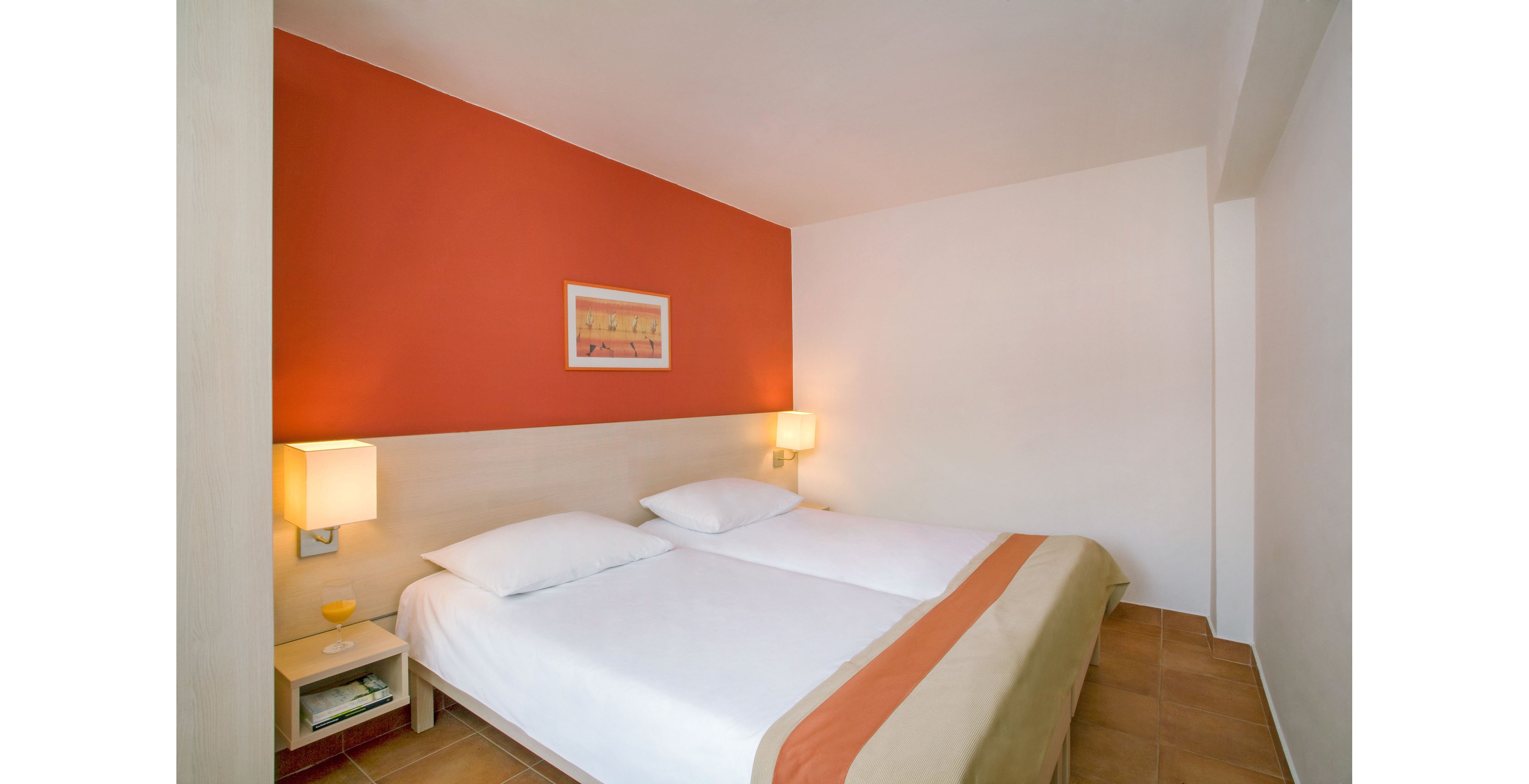 A clean, minimalist hotel bedroom features two neatly made twin beds pushed together with white linens and a beige and orange runner. The room has a warm orange accent wall behind the headboard, contrasting with the otherwise white walls. Two modern bedside lamps cast a soft, cozy light on the wooden nightstands. A framed artwork hangs above the beds, adding a subtle decorative touch to the simple interior.