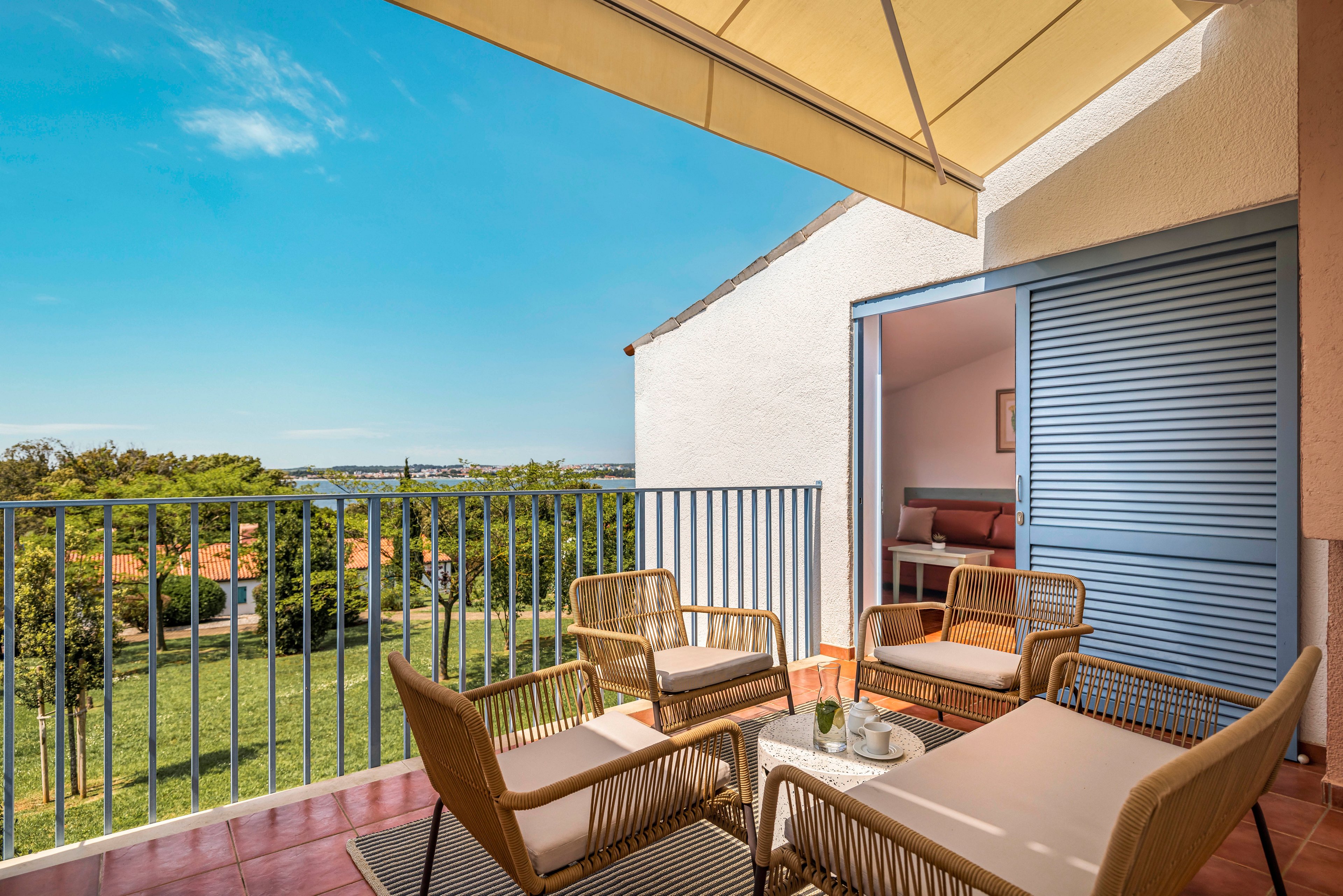 A bright balcony terrace features a set of wicker chairs and cushioned benches arranged around a small coffee table. The space opens from an interior living room through blue sliding shutters, creating an inviting indoor-outdoor connection. A large awning provides shade while the terrace overlooks a green lawn, trees, and distant rooftops under a clear blue sky. The scene suggests a relaxed, vacation-like atmosphere ideal for lounging or casual conversation.