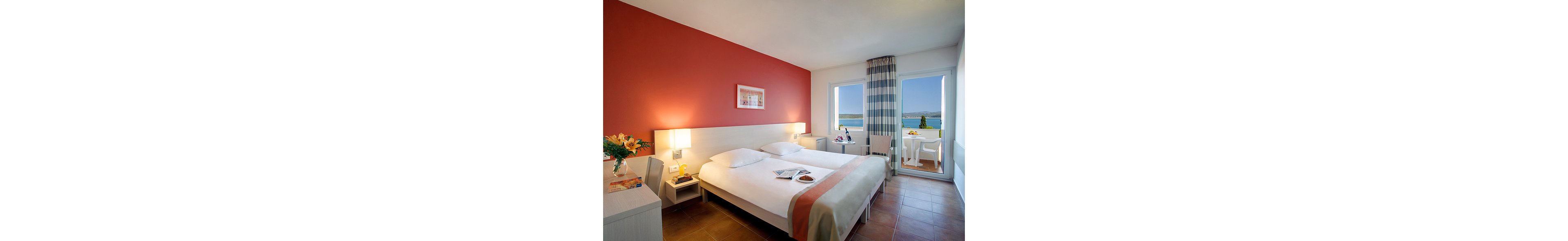 A bright hotel room features a neatly made double bed with white linens and a soft coral accent runner. The space includes bedside tables with warm lamps, a small desk, and a chair positioned near a large window. A balcony door opens to an outdoor seating area with a table and chairs overlooking the sea and distant shoreline. Fresh flowers, magazines, and a light snack on the bed add a welcoming, relaxed atmosphere.