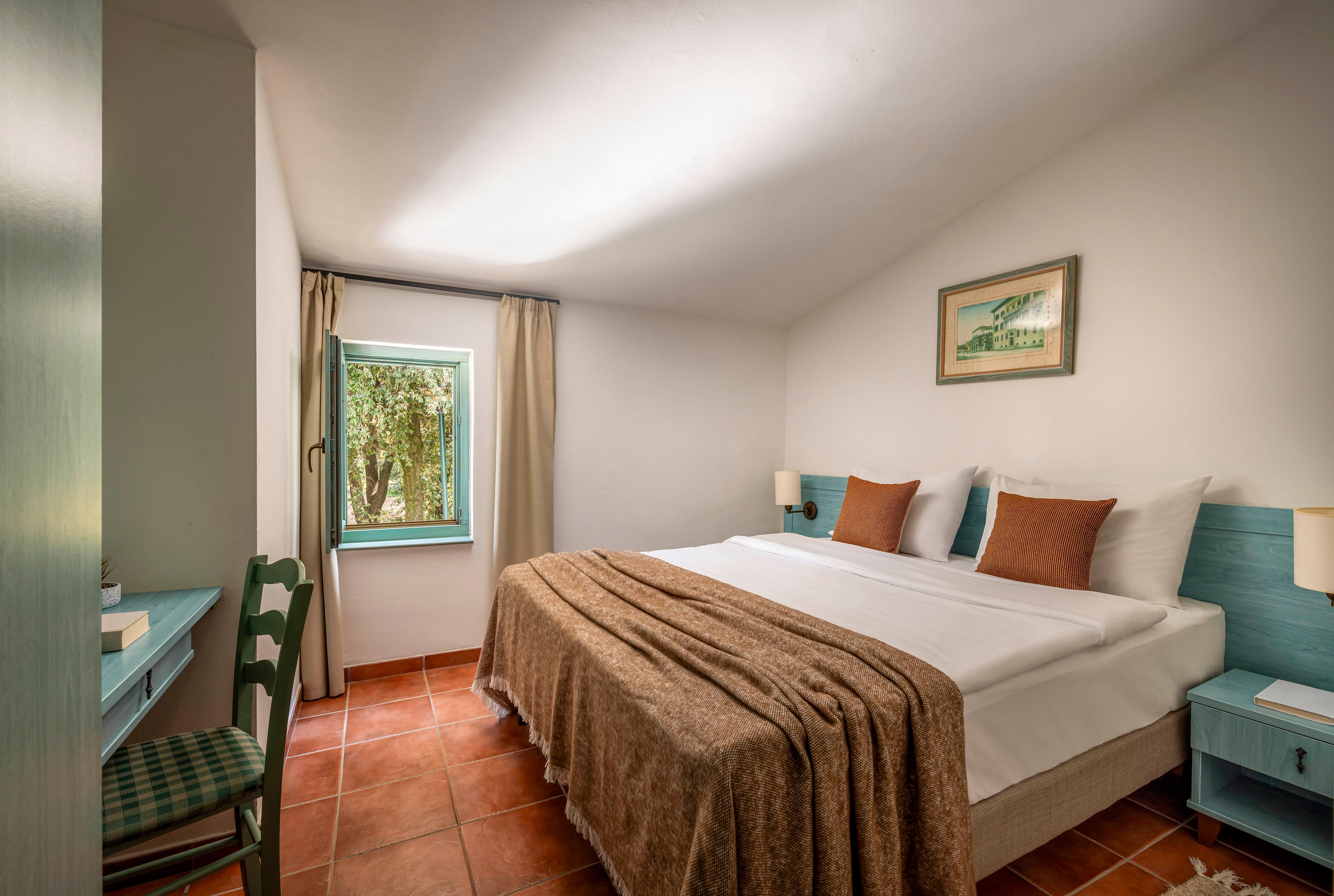 A neatly made double bed with white linens and brown throw blankets is the focal point of this cozy bedroom. The room features terracotta tile flooring, light-colored walls, and teal bedside tables with matching lamps. A small desk and chair sit near a single window that lets in natural light and offers a view of greenery outside. The overall atmosphere is calm and inviting, suitable for a hotel or guest room stay.