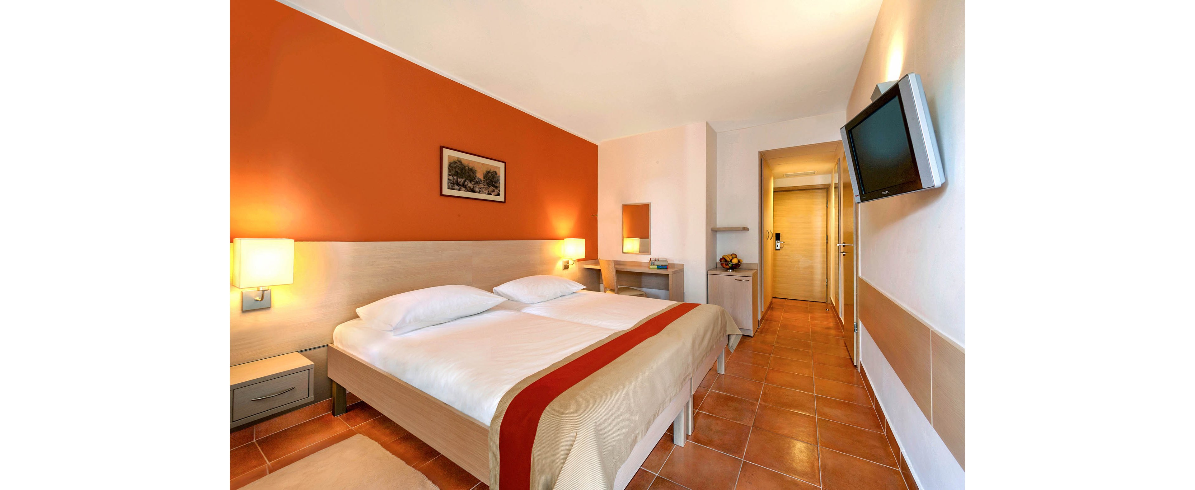 A contemporary hotel room features two neatly made twin beds with white linens and a beige duvet accented by a red runner. The space includes warm orange and neutral walls, bedside tables with lit lamps, and a small desk with a mirror. A flat-screen television is mounted on the wall opposite the beds, and the floor is covered with terracotta-colored tiles. The room leads to a hallway with a door, suggesting an entrance or bathroom area.