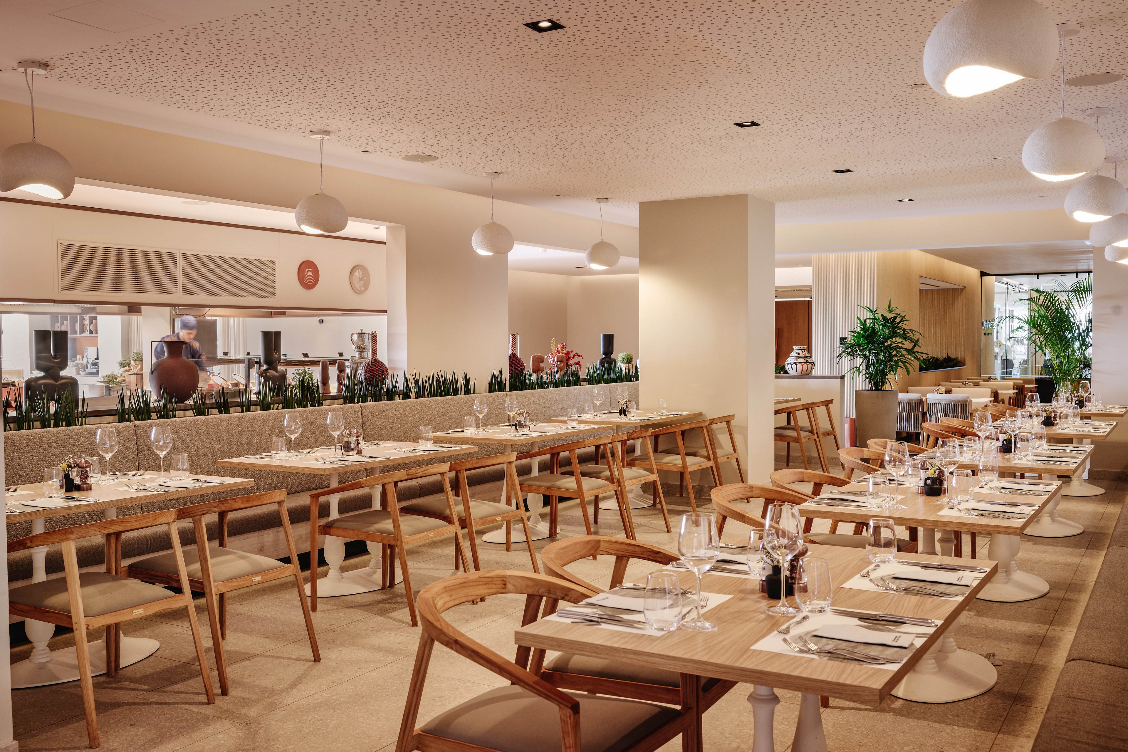 Contemporary restaurant dining area featuring light wooden tables and chairs is shown set for service. Wine glasses, cutlery, and napkins are neatly arranged on each table, with small plants and condiments adding detail. An open kitchen with staff working is visible in the background, separated by a low partition with greenery. Warm lighting, neutral tones, and clean lines create a relaxed, upscale atmosphere.
