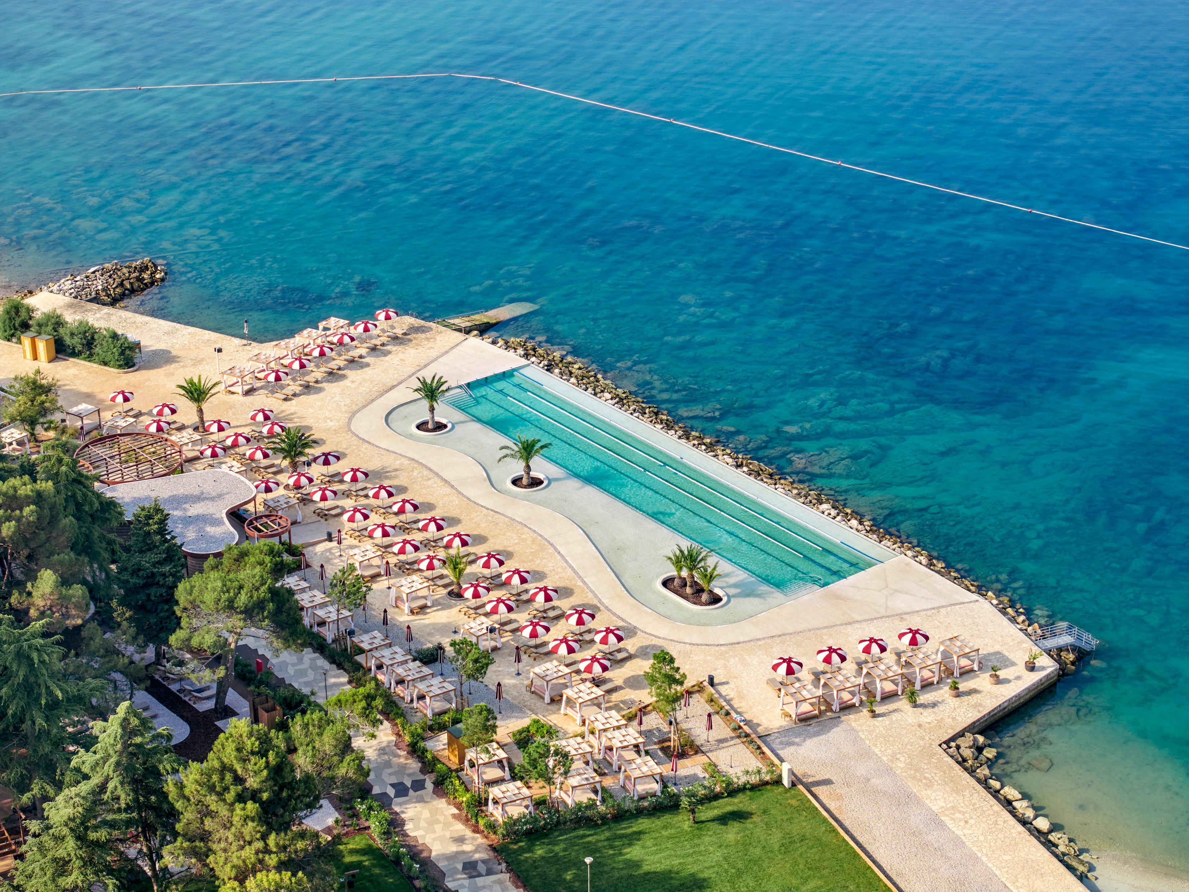Aerial view of a coastal resort featuring a long rectangular swimming pool beside clear blue sea water. Numerous red and white striped umbrellas shade sun loungers arranged in neat rows along the stone terrace. Palm trees and landscaped greenery frame the leisure area, creating a relaxed vacation atmosphere. The scene suggests a tranquil, sunny day at an upscale beachfront destination.