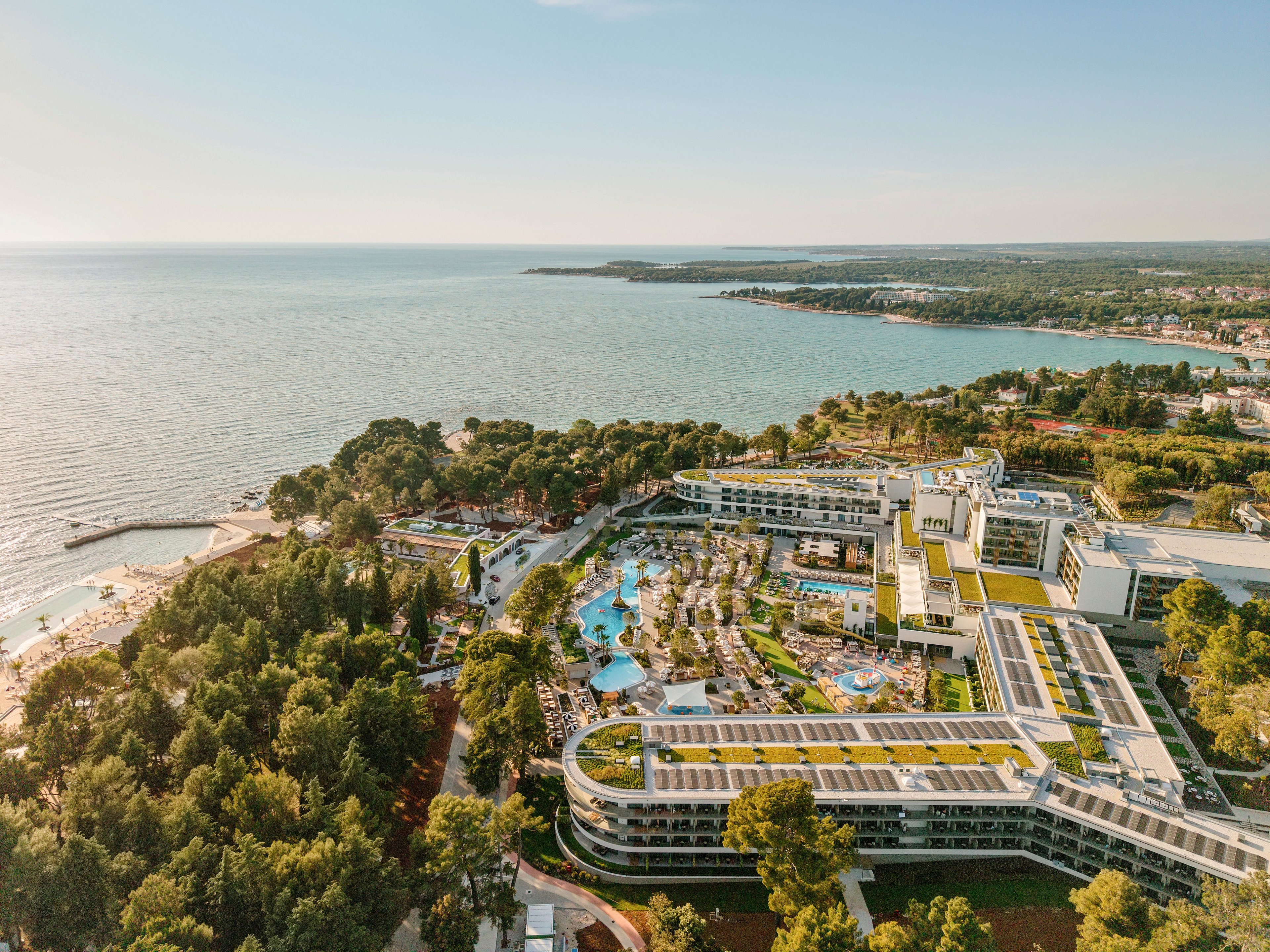 A large contemporary resort complex is captured from above along a scenic coastline. Multiple white buildings with terraces surround turquoise swimming pools and landscaped gardens. The resort is bordered by dense green trees and opens onto sandy beaches beside a calm sea. The wider shoreline and nearby town stretch into the distance under warm, clear light, suggesting a relaxed holiday atmosphere.