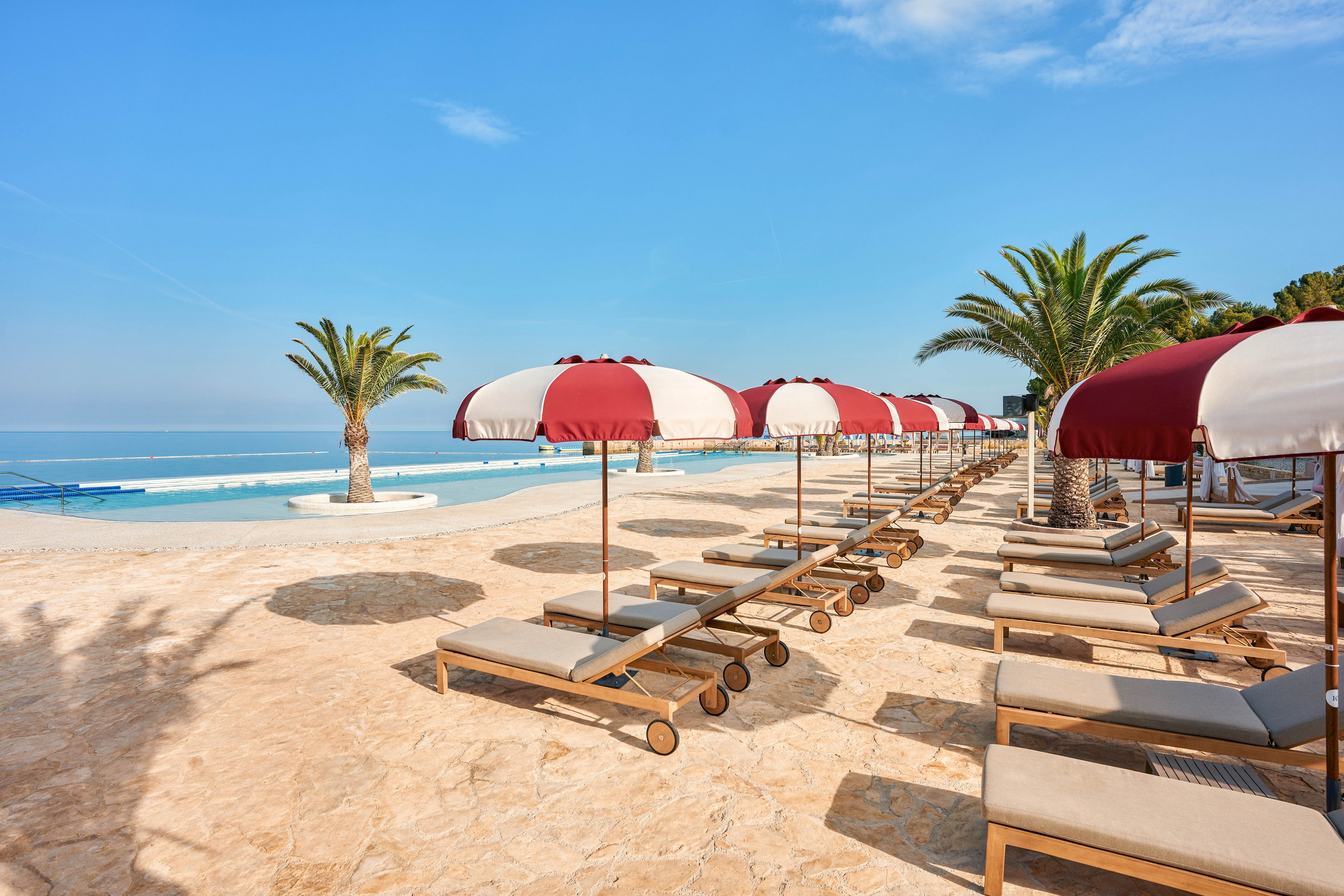 A row of cushioned lounge chairs with red and white striped umbrellas lines a sunlit terrace beside a large infinity-style pool. The setting overlooks a calm blue sea under a clear sky, creating a tranquil coastal atmosphere. Palm trees add a tropical touch to the scene, and the area appears quiet and empty, ready for relaxing guests. Warm stone flooring and soft neutral tones contrast with the bright blue water and sky.