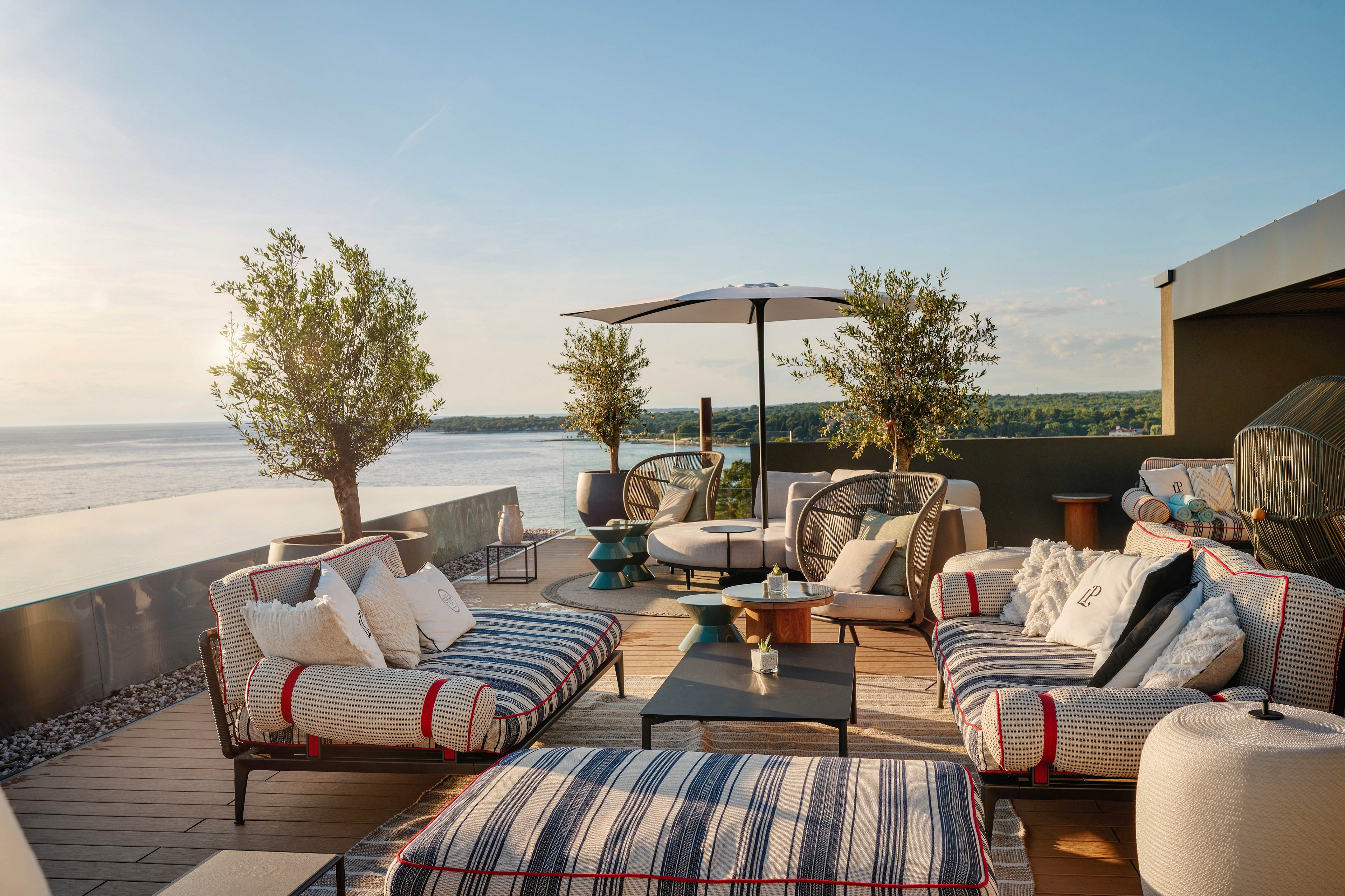 A contemporary rooftop terrace features cushioned lounge seating and a central coffee table under a large umbrella. Striped sofas and accent chairs are arranged for socializing, with decorative pillows and small side tables adding comfort. Potted trees and plants frame the space, while a glass railing opens to a wide view of the water and distant shoreline. Warm evening light creates a relaxed, inviting atmosphere ideal for outdoor entertaining.