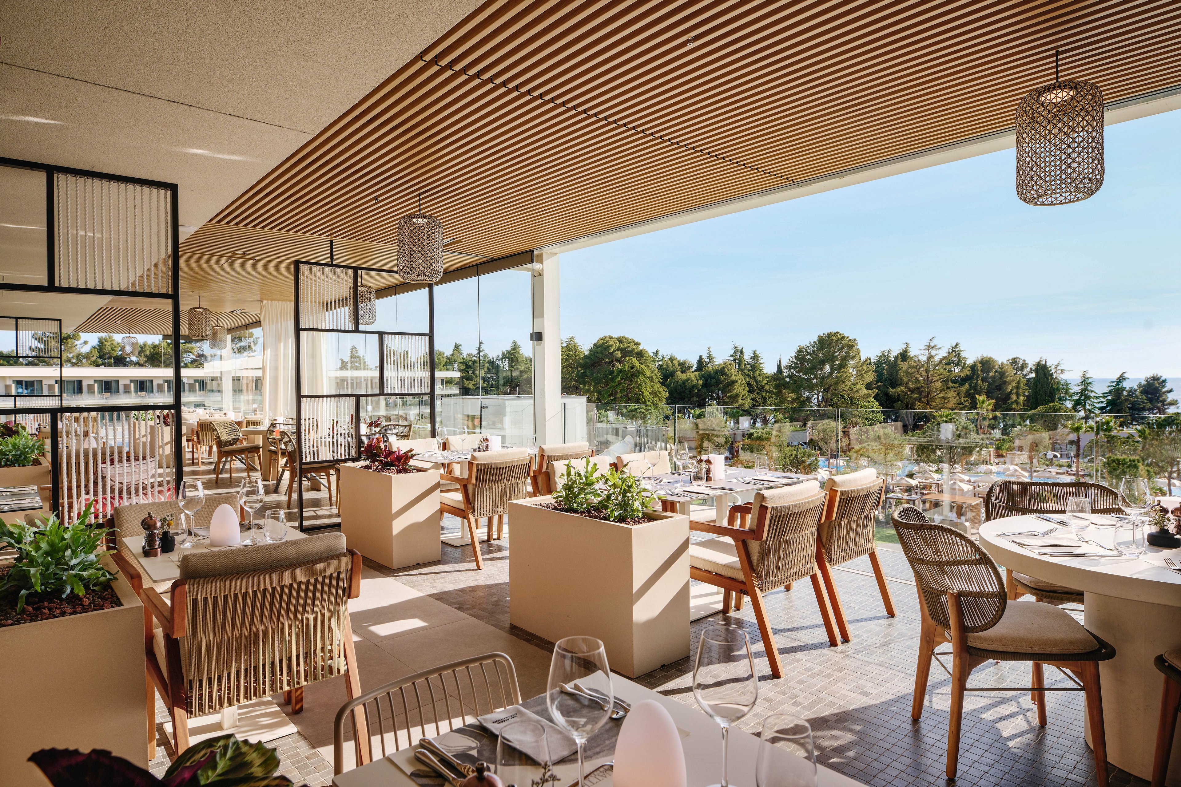 A contemporary restaurant terrace features neatly arranged tables and wicker chairs under a slatted wooden ceiling. The setting overlooks a sunny outdoor area with greenery and distant buildings, creating an inviting atmosphere. Table settings include glassware, plates, and decorative centerpieces, suggesting readiness for guests. Warm natural light and neutral tones contribute to a relaxed yet upscale dining experience.