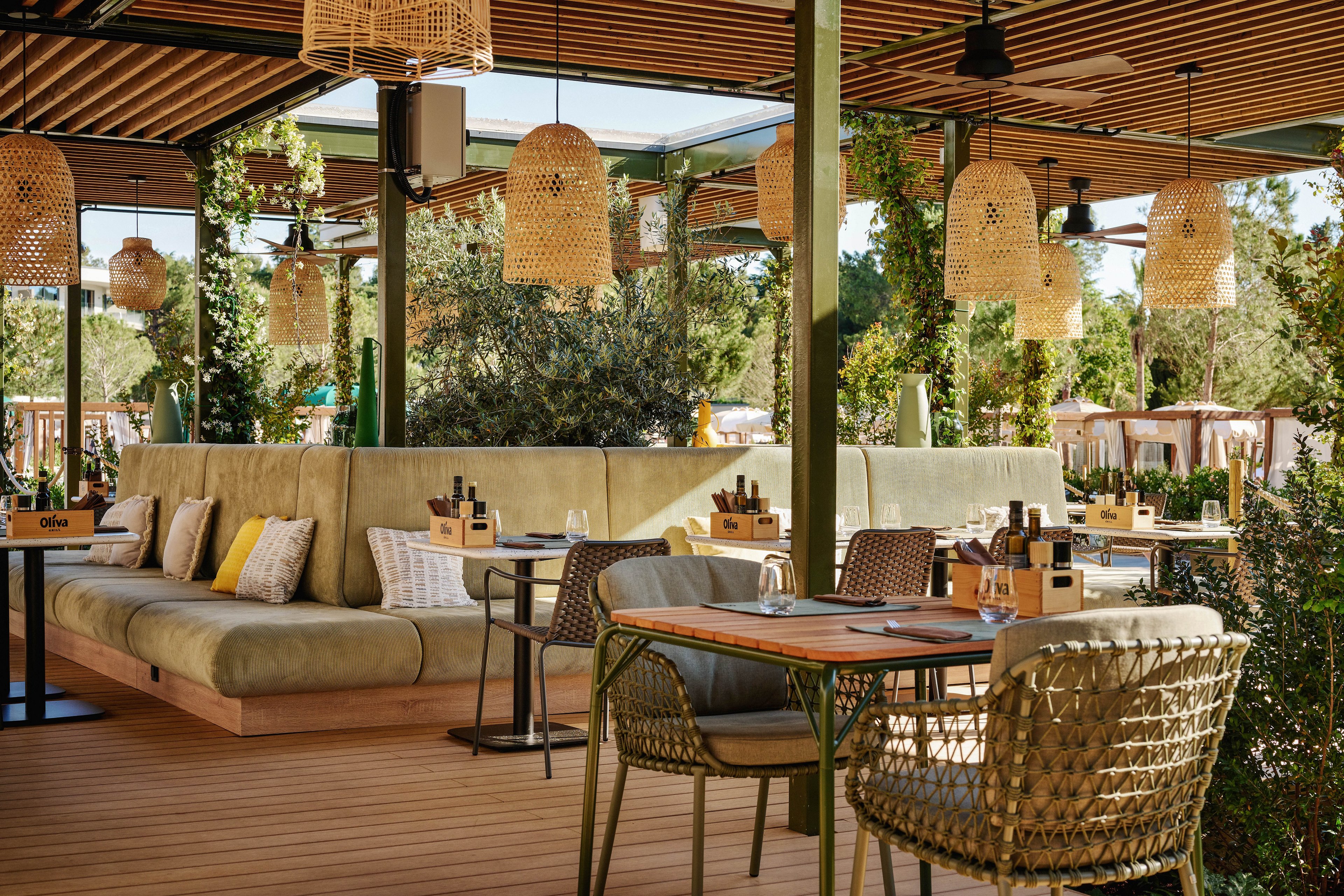 An inviting outdoor restaurant terrace features cushioned bench seating and woven wicker chairs around wooden tables. Warm pendant lights hang from the ceiling, complementing the natural wood floor and green metal structure. Small table caddies hold condiments and bottles, while decorative pillows add comfort and color. Lush greenery and trees surround the space, creating a relaxed, contemporary dining atmosphere.