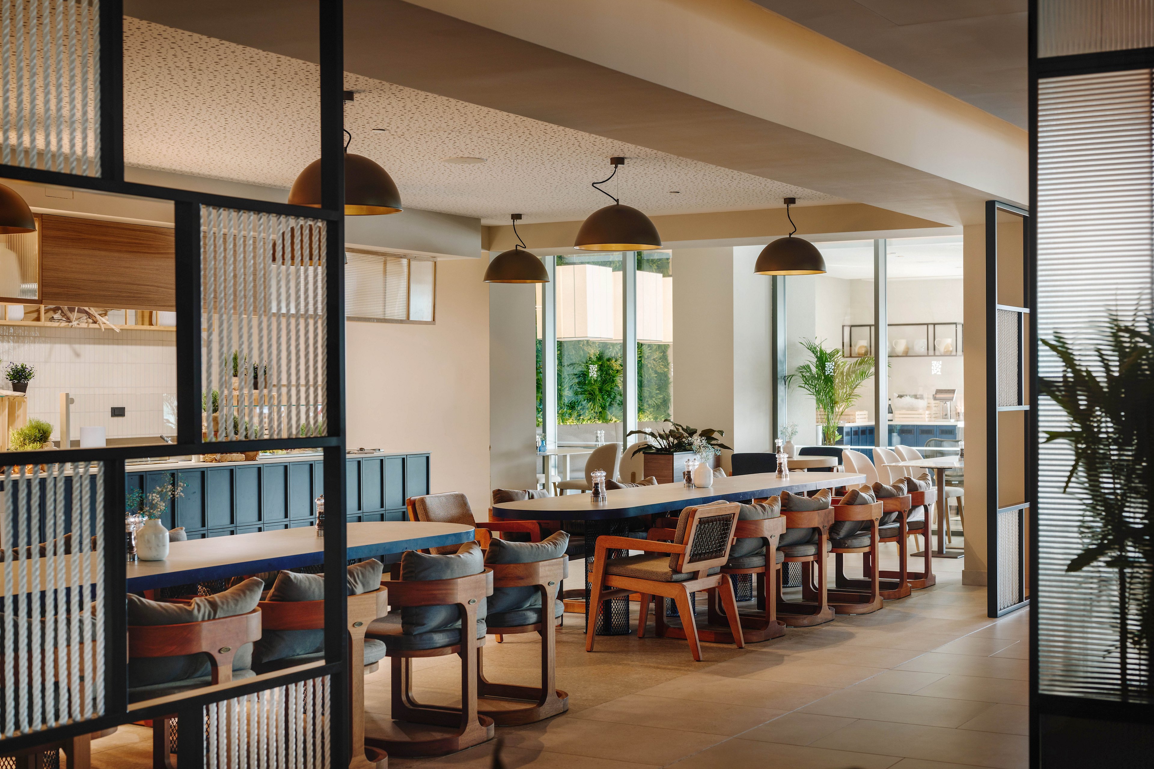 Contemporary restaurant dining area featuring long communal tables and cushioned chairs is shown in warm natural light. Large windows and glass partitions create an open, airy atmosphere with views to the outside. Pendant lights hang above the tables, and potted plants add greenery throughout the space. The overall design feels clean, inviting, and suitable for casual or business dining.