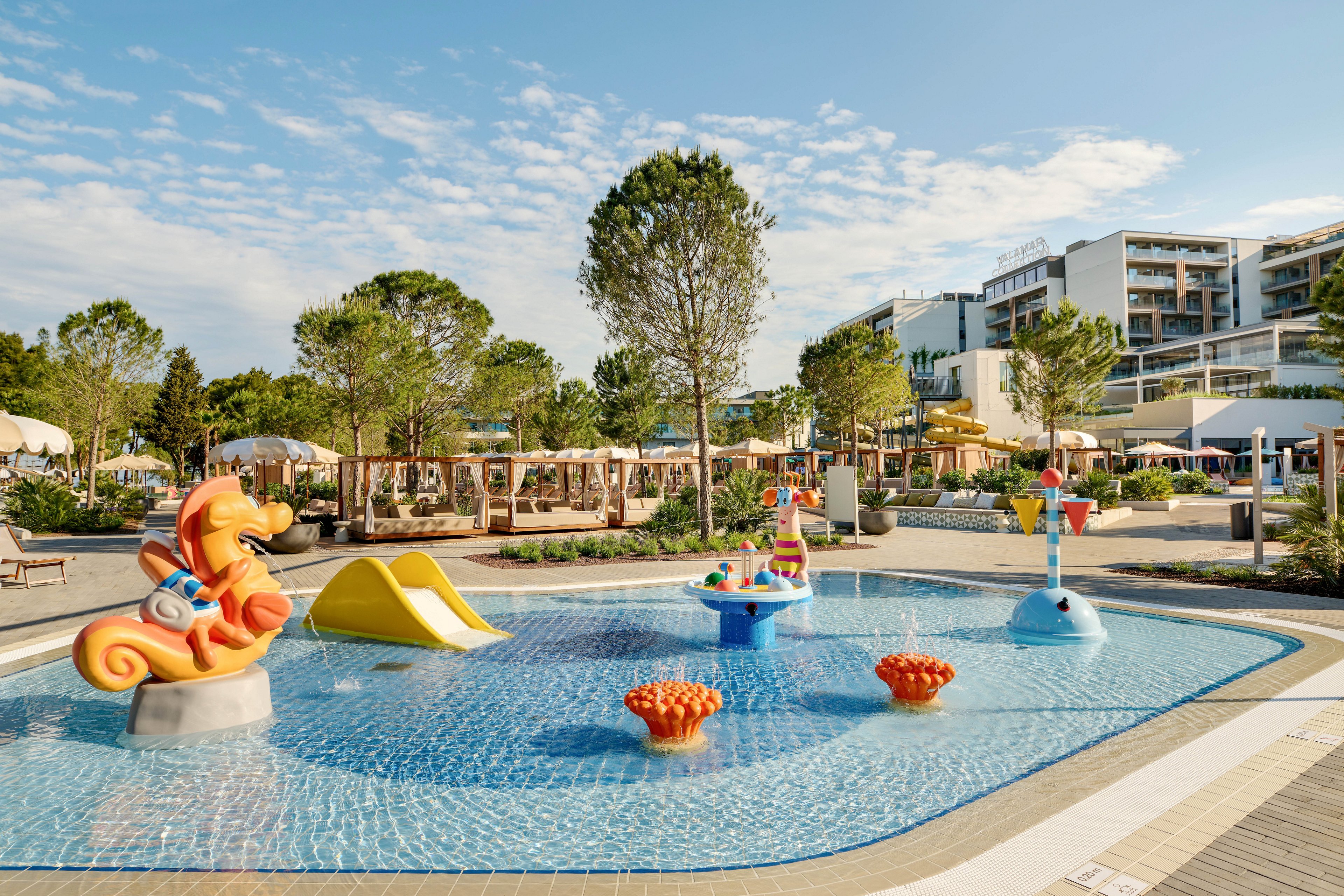 Outdoor children’s splash pool with colorful water play structures is shown at a modern resort. The pool features a seahorse slide, small yellow slide, and interactive fountains in shallow water. Sun loungers and cabanas line the background beneath tall green trees. A contemporary multi-story hotel building rises behind the pool under a bright blue sky with scattered clouds.