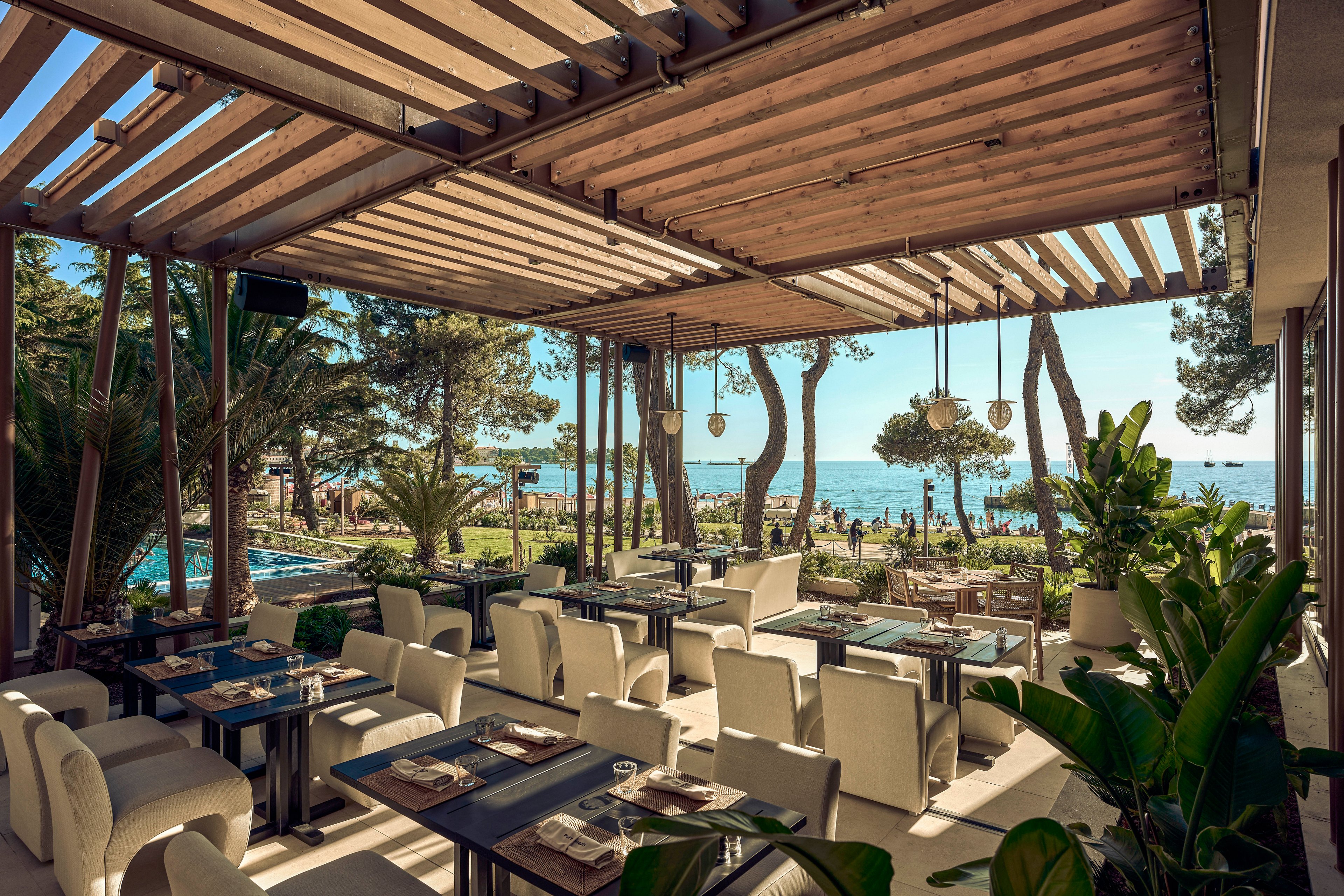 An elegant outdoor restaurant terrace overlooks a lush garden and the sea beyond. Neatly arranged tables and cushioned chairs sit under a wooden pergola with hanging lights. Palm trees, greenery, and a distant pool area create a resort-like atmosphere. The scene suggests a relaxed dining experience with scenic coastal views on a sunny day.