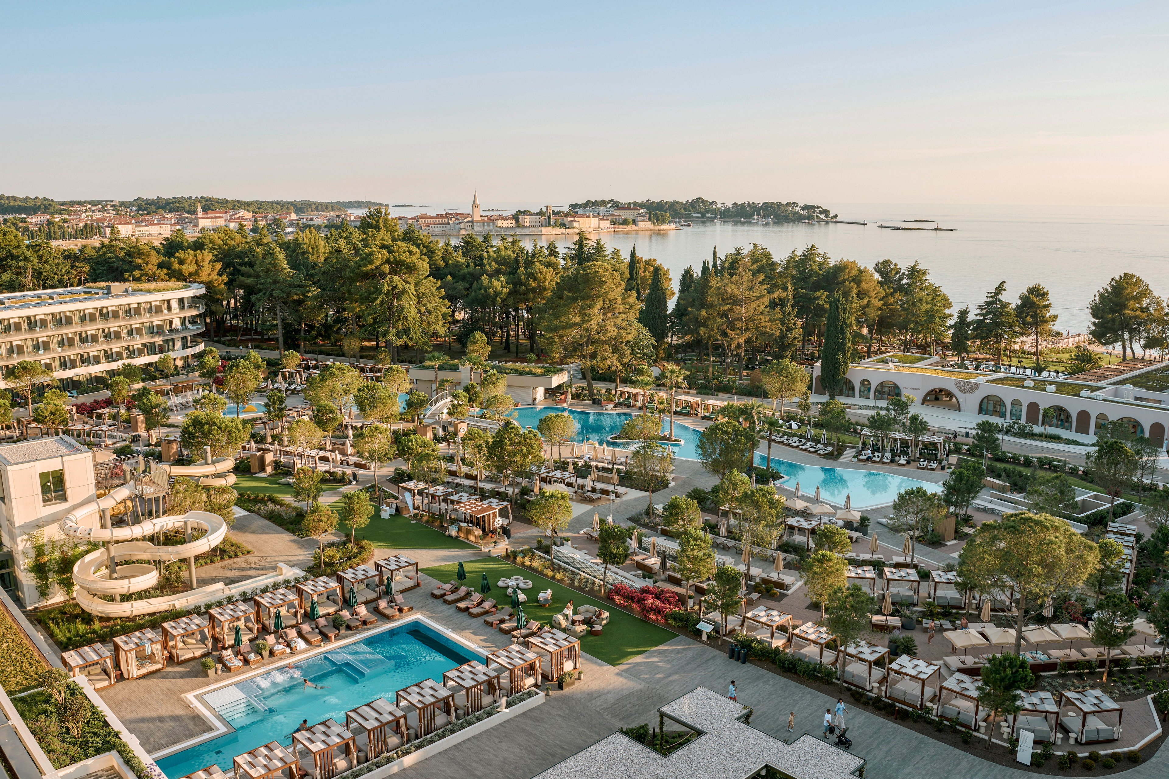A large upscale resort complex features multiple outdoor swimming pools surrounded by sun loungers and cabanas. Modern hotel buildings and landscaped gardens fill the foreground, while the coastline and calm sea stretch into the distance. The warm sunset light creates a tranquil atmosphere over the entire property. Trees and pathways weave through the scene, emphasizing the expansive, well-planned vacation destination.