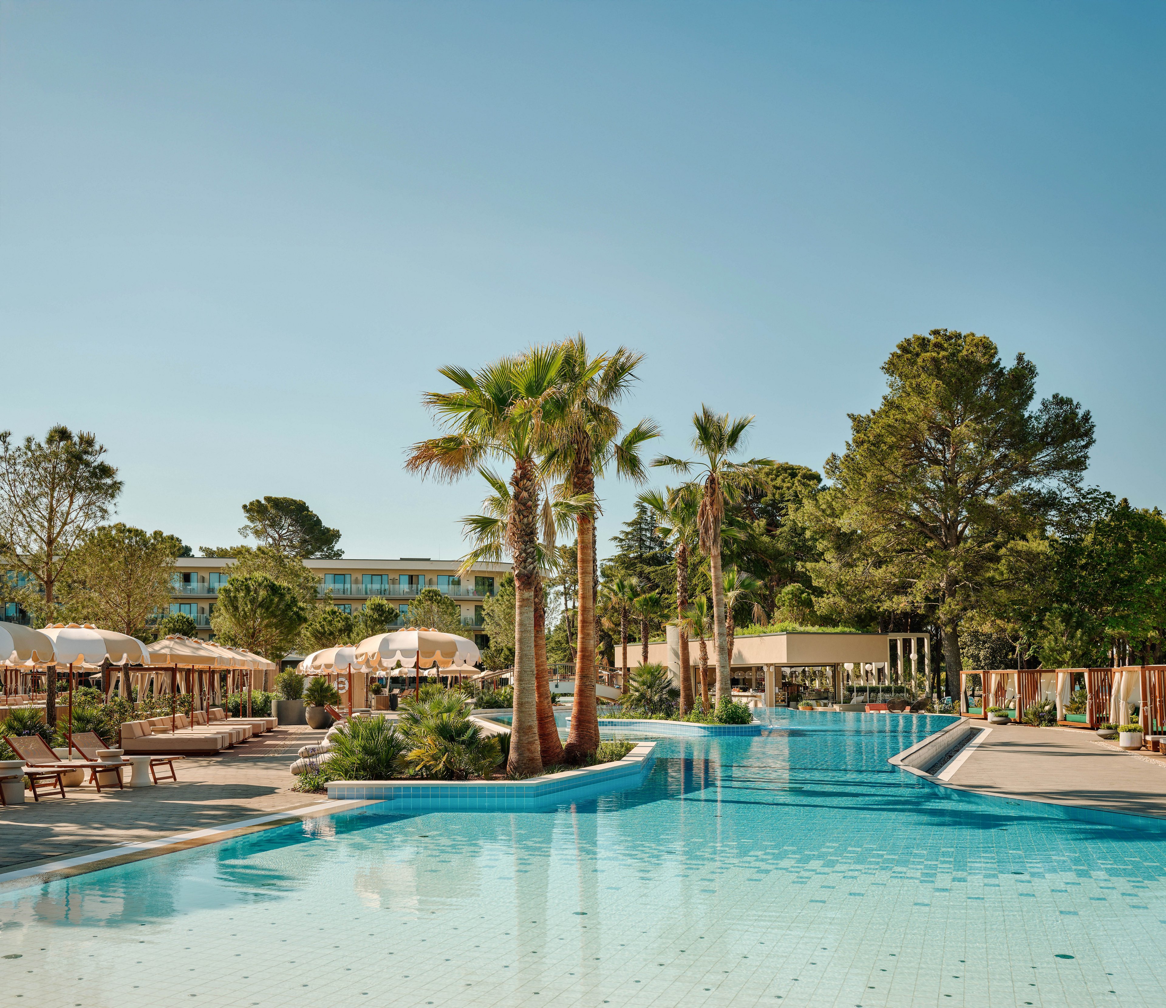 A large outdoor swimming pool stretches across a sunny resort, bordered by tall palm trees and lush greenery. Lounge chairs and umbrellas line the poolside, creating a relaxed vacation atmosphere. In the background, low-rise hotel buildings and cabana-style structures are visible under a clear blue sky. The scene suggests a tranquil holiday destination focused on leisure and relaxation.