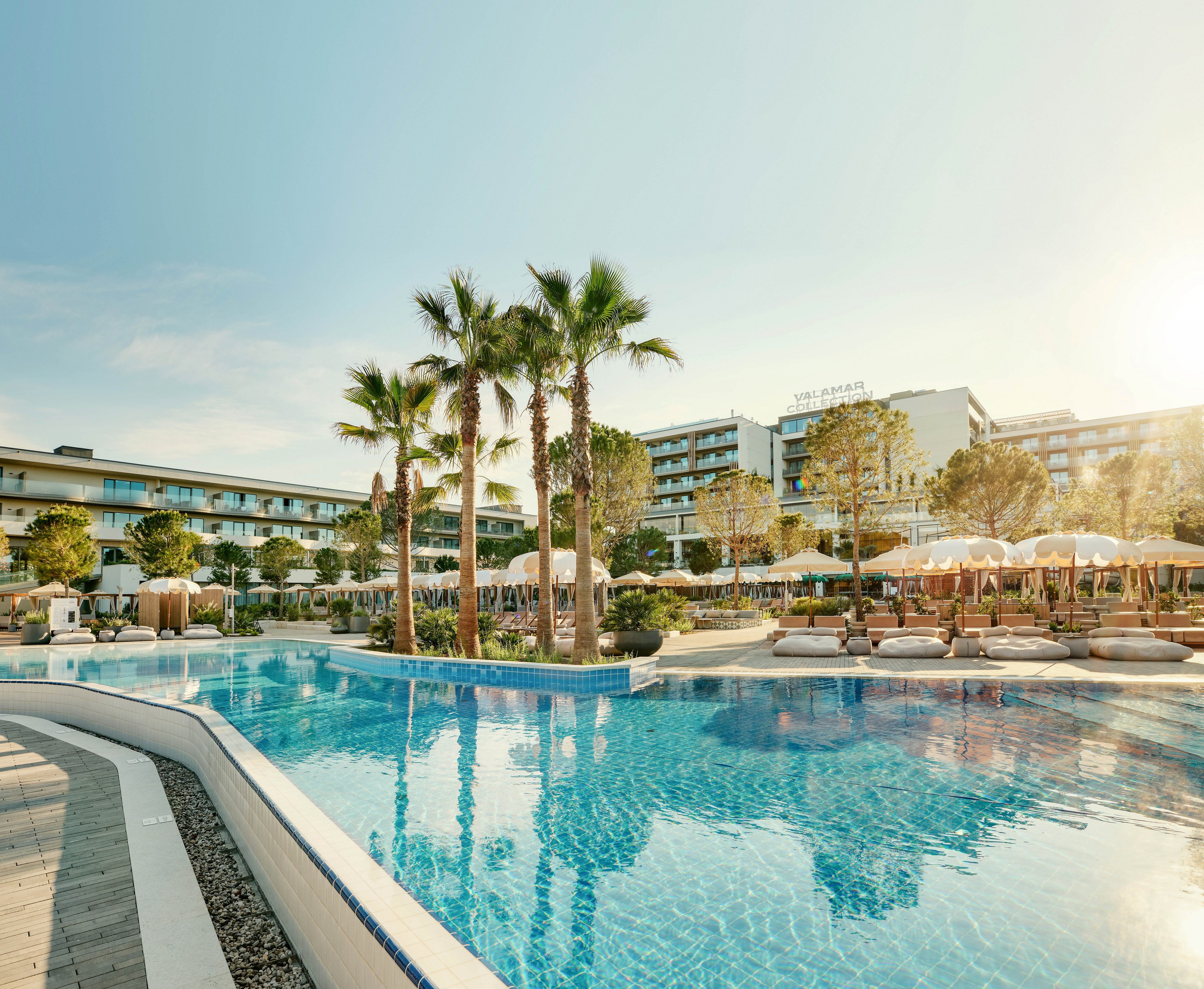 A large outdoor swimming pool reflects palm trees and modern resort buildings in the soft light of sunrise or sunset. Sun loungers and parasols line the poolside, suggesting a relaxing vacation atmosphere. The architecture is contemporary, with multiple stories and balconies overlooking the water. The overall scene feels tranquil and inviting, with warm tones and clear blue water.