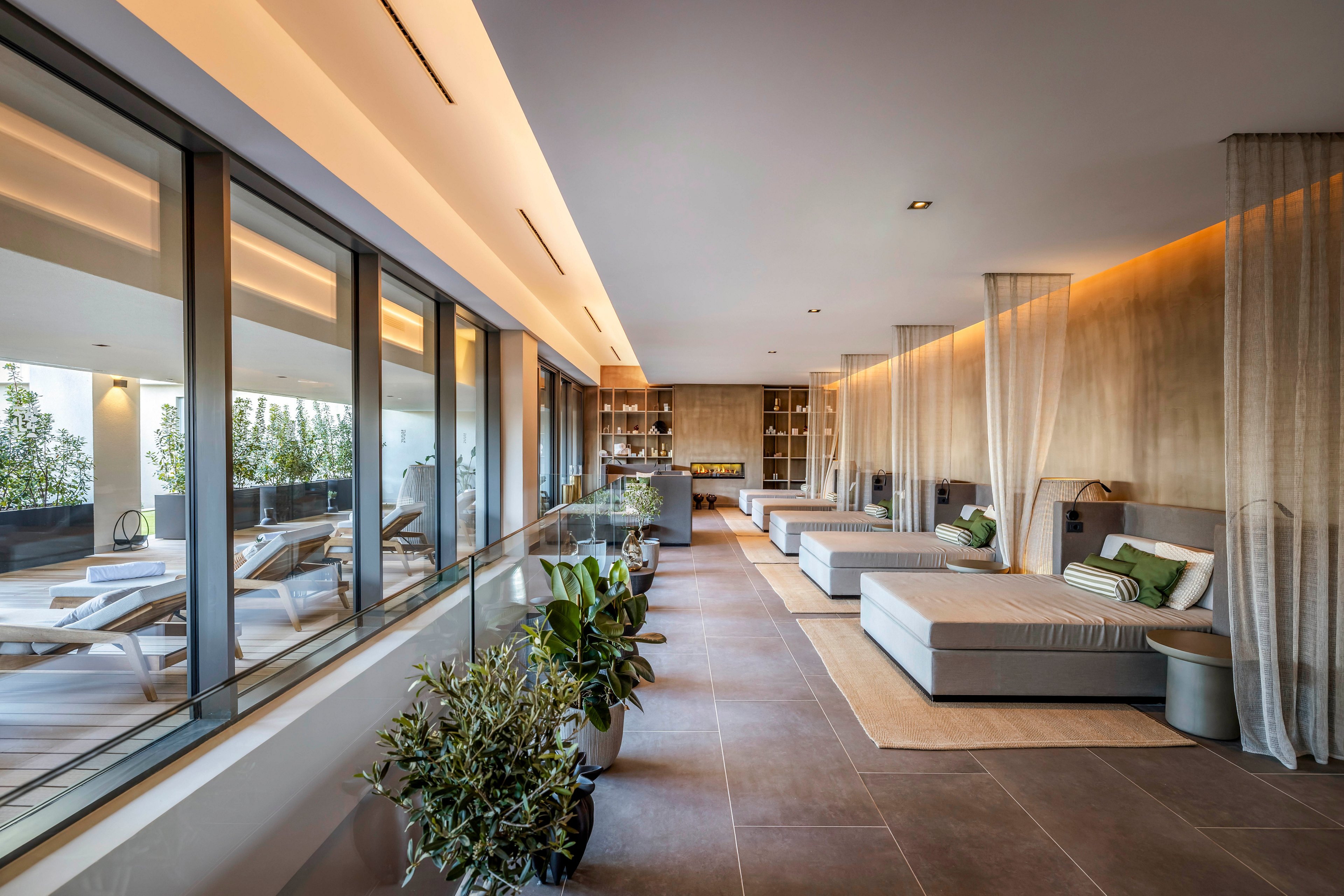 A contemporary spa lounge features a row of cushioned daybeds with pillows and throws. Floor-to-ceiling windows separate the relaxation area from an adjacent terrace with sun loungers. Warm indirect lighting, wood paneling, and neutral tones create a calm, luxurious atmosphere. Potted plants and sheer curtains add softness and a natural touch to the minimalist interior.