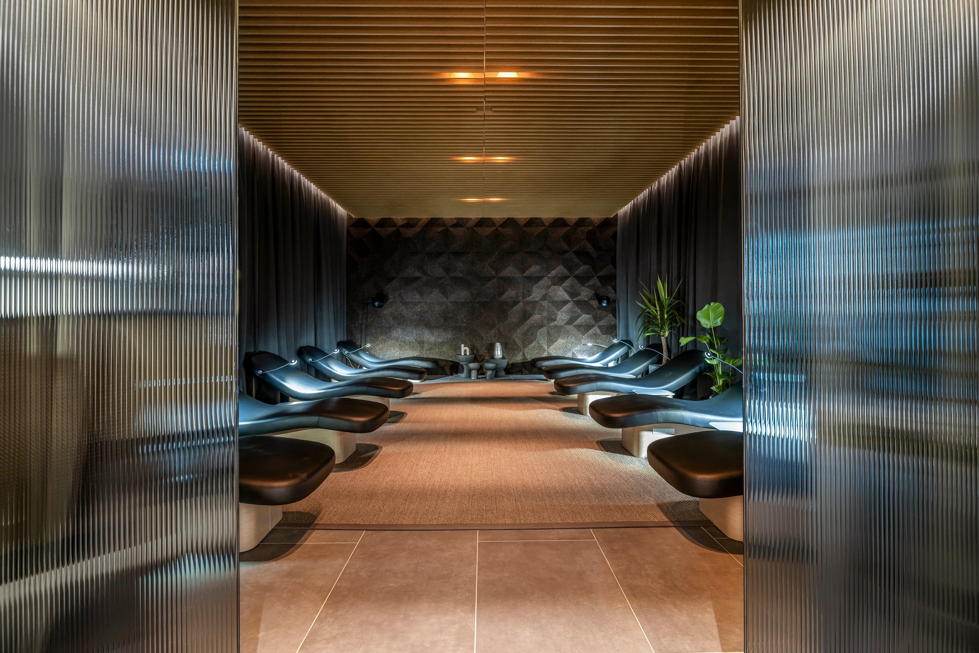 A contemporary relaxation lounge features a row of sleek black recliner chairs on both sides of a central aisle. The space is enclosed by textured walls and curtains, leading the eye toward a small seating area at the back. Warm ceiling lights and soft floor lighting create a calm, intimate atmosphere. Minimalist decor and a few green plants add a touch of nature to the modern interior design.