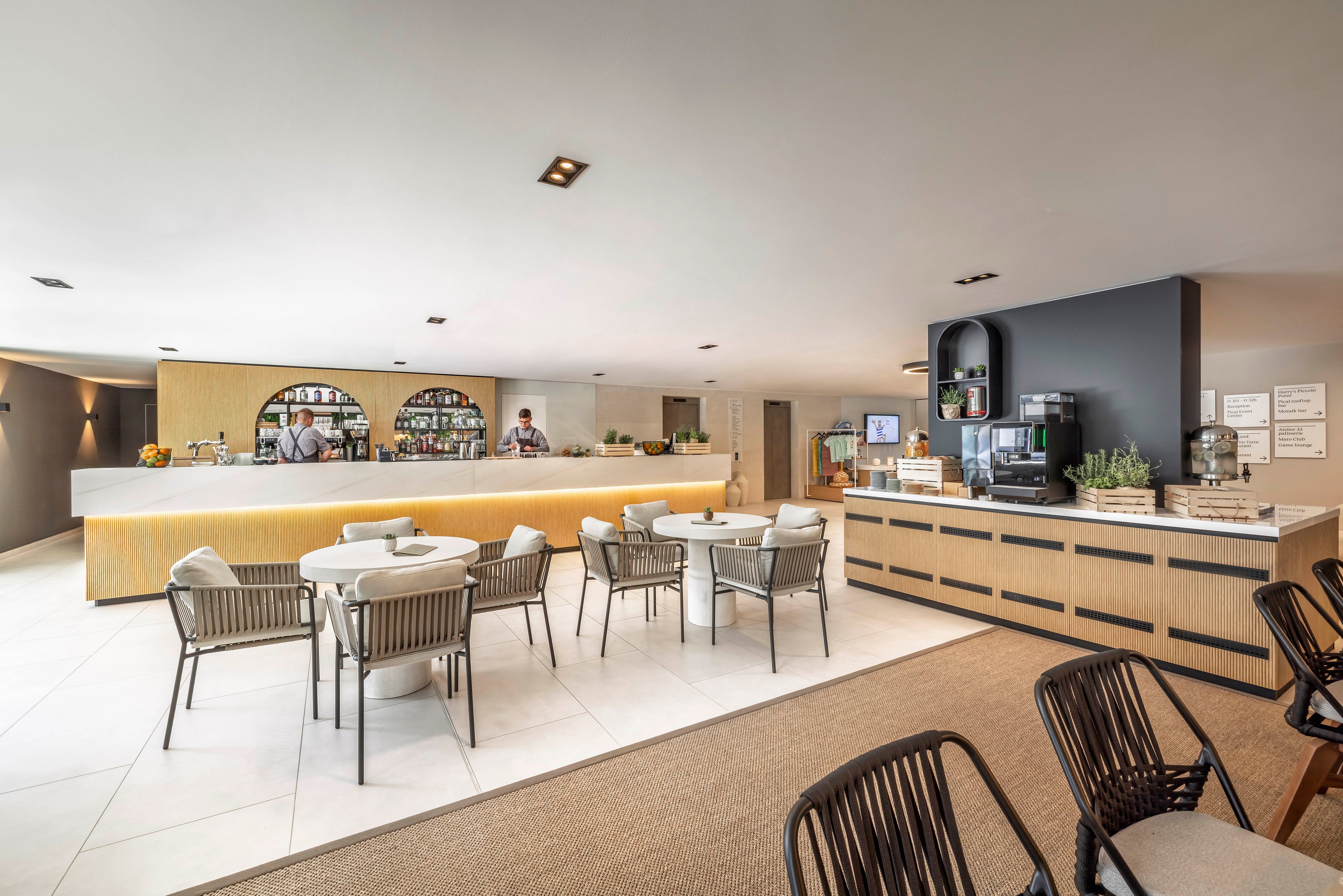 Spacious contemporary lounge area featuring a long bar counter and a separate self-service cafe station. Round tables with cushioned chairs are arranged on a light tiled floor, while darker chairs sit on a carpeted section in the foreground. The bar displays bottles and glassware on arched shelves, and the cafe counter holds a coffee machine, plants, and neatly arranged items. Warm neutral tones and clean lines create a relaxed yet upscale atmosphere suitable for socializing or casual meetings.