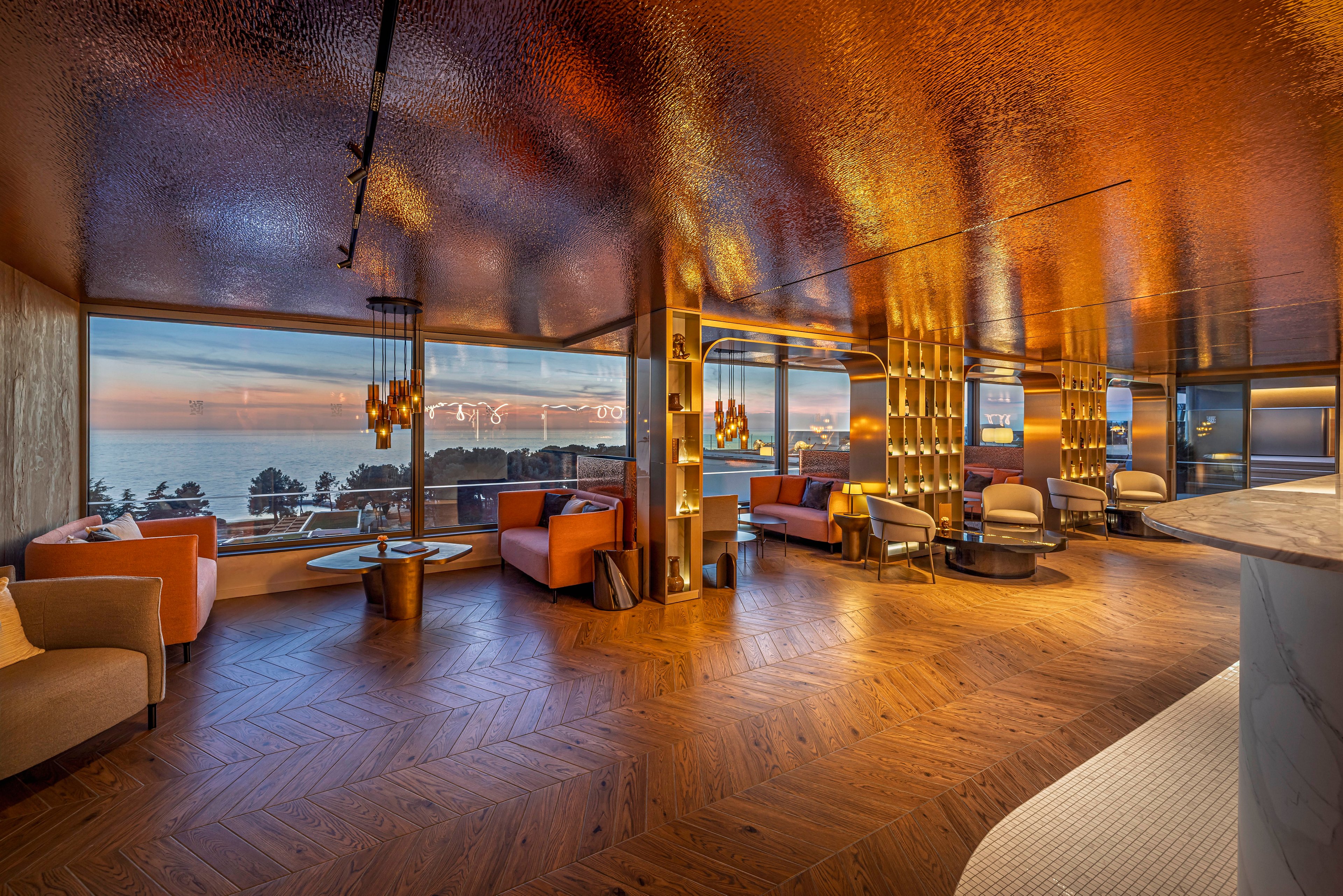 A spacious luxury lounge features contemporary seating arranged around large floor-to-ceiling windows overlooking the ocean at sunset. Warm golden lighting reflects off metallic surfaces and a textured ceiling, creating a rich, inviting atmosphere. Modern armchairs and sofas in muted orange and beige tones sit on a patterned wooden floor. The scene conveys an upscale, relaxing environment with dramatic coastal views and sophisticated interior design.