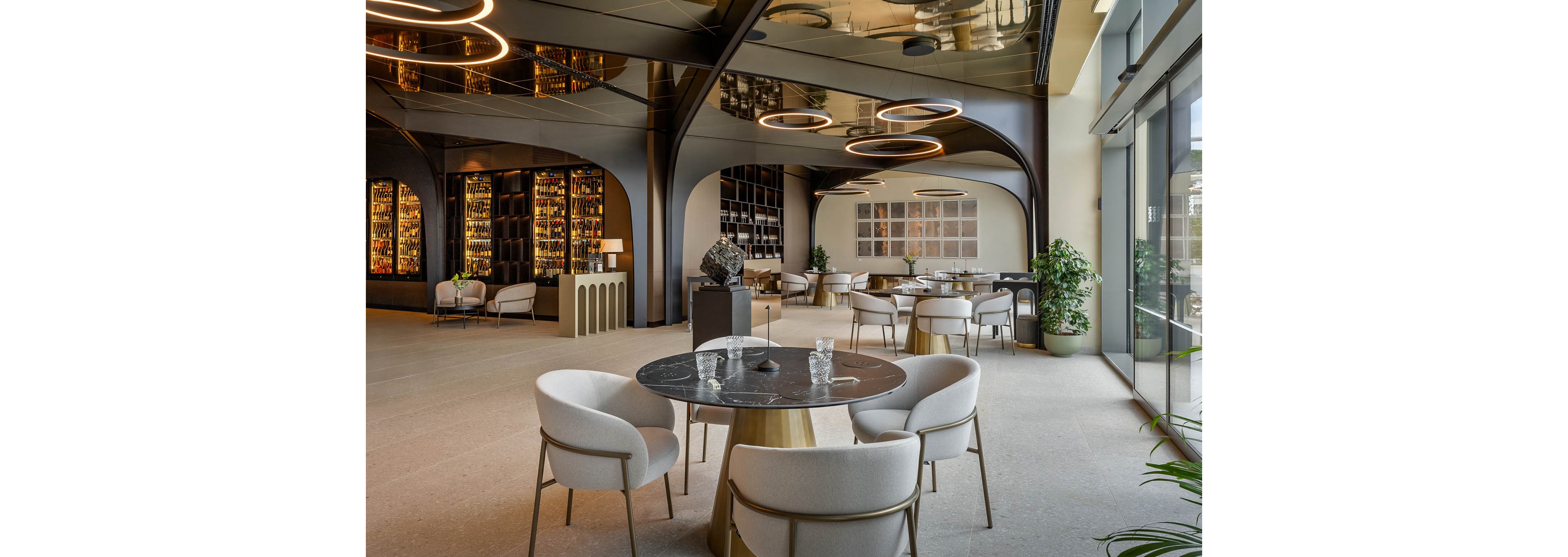 A contemporary lounge area features round black marble tables surrounded by plush white chairs. The setting is spacious with large windows, modern circular ceiling lights, and a wall of illuminated shelves displaying bottles. The atmosphere is sophisticated and inviting, with neutral tones and greenery adding to the upscale ambiance. No visible text or numeric values are present in the image.