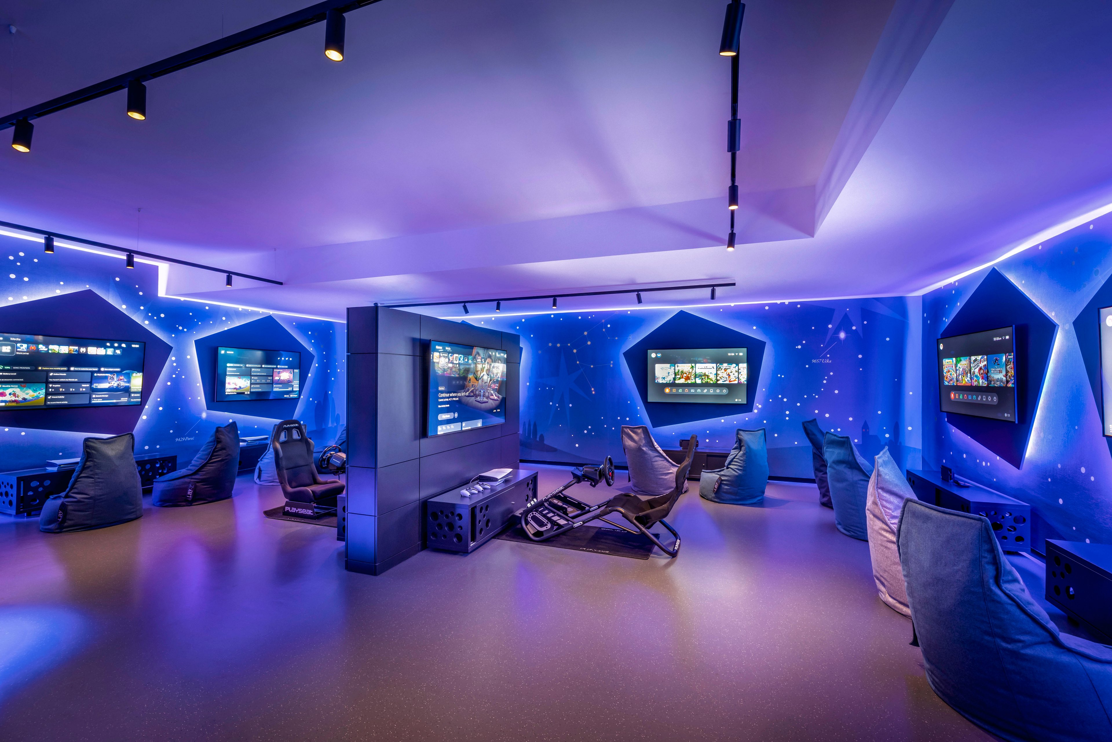 A futuristic gaming lounge features multiple large screens and racing simulator chairs, surrounded by bean bags for comfortable seating. The room is illuminated with vibrant blue and purple LED lights, creating an energetic and immersive atmosphere. The walls display digital visuals and gaming interfaces, enhancing the high-tech ambiance. No visible text or numeric values are present in the image.