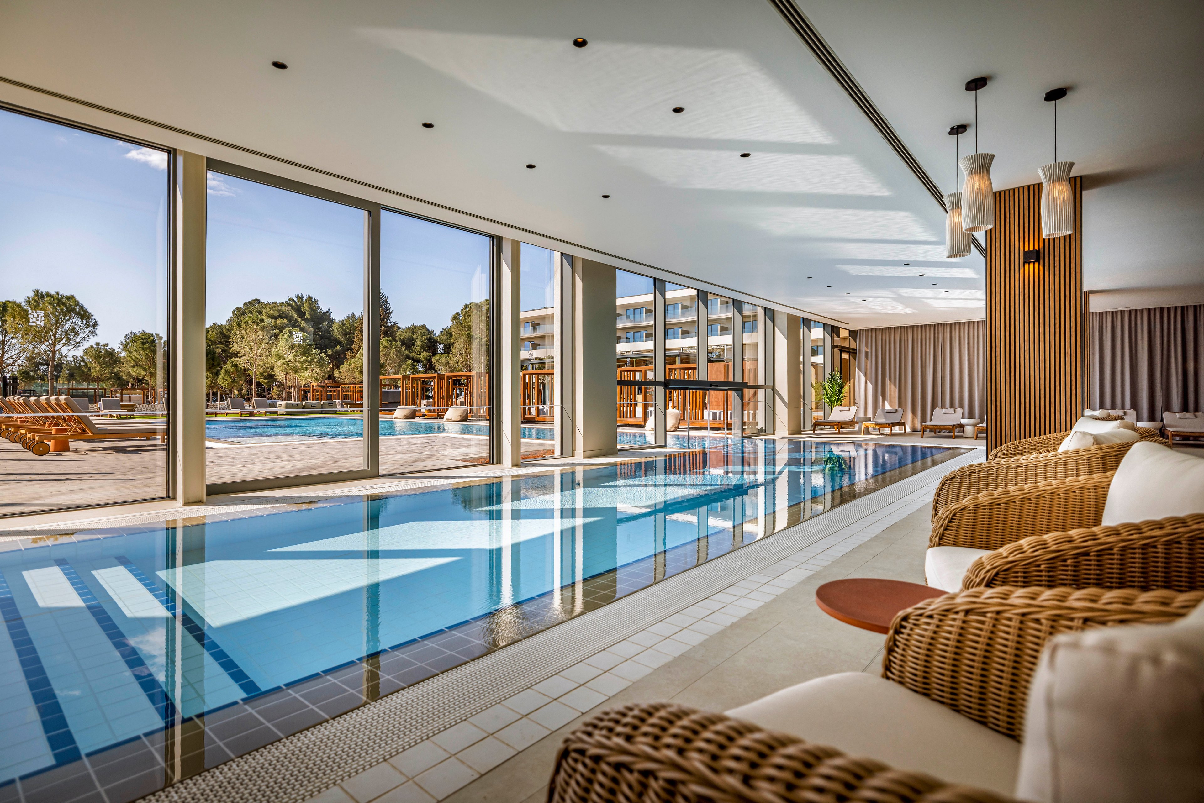 The image features a stylish indoor swimming pool area with wicker lounge chairs and plush cushions. Large floor-to-ceiling windows provide a clear view of an outdoor pool and landscaped surroundings, including trees and modern buildings. The setting is bright and inviting, with natural light enhancing the warm, neutral palette and contemporary decor. No visible text or numeric values are present in the image.