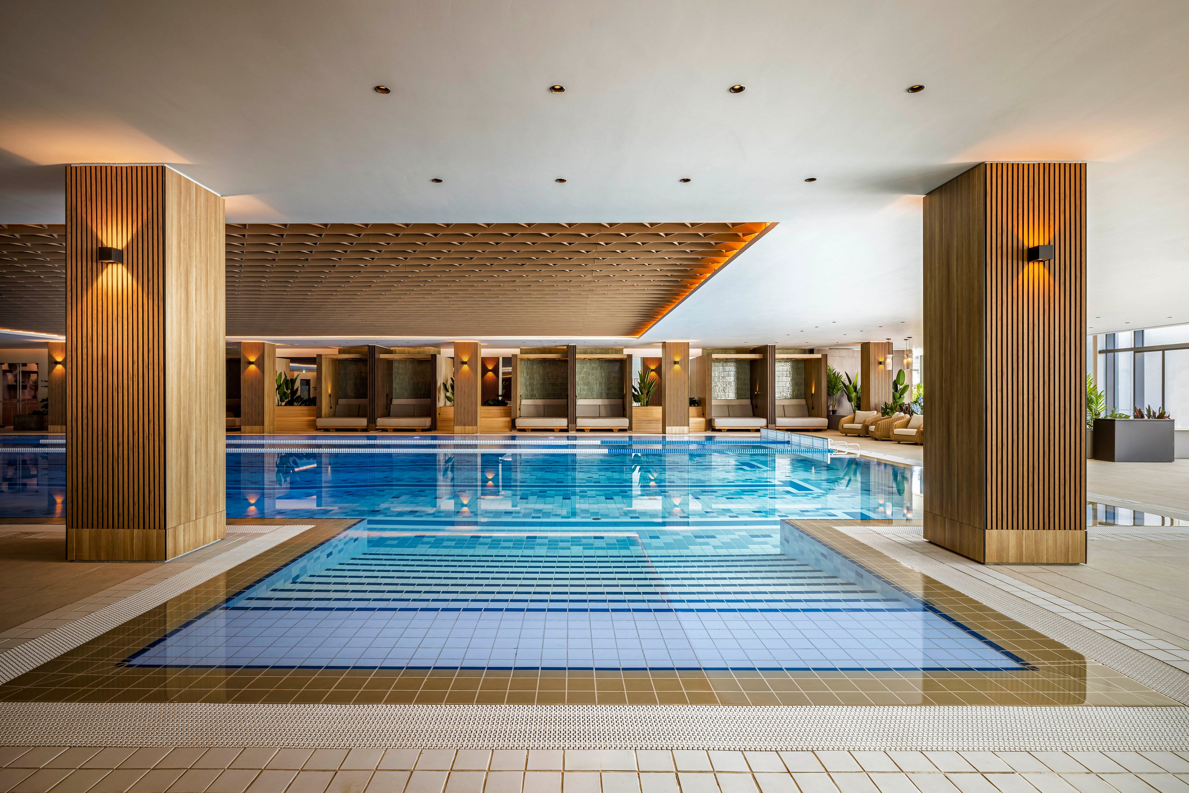 A spacious indoor swimming pool is featured in a contemporary setting with wooden columns and recessed ceiling lights. The pool area includes a shallow entry section and several private lounge cabanas along the far wall. The scene is well-lit, with clean lines and a calm, inviting atmosphere, emphasizing relaxation and luxury.