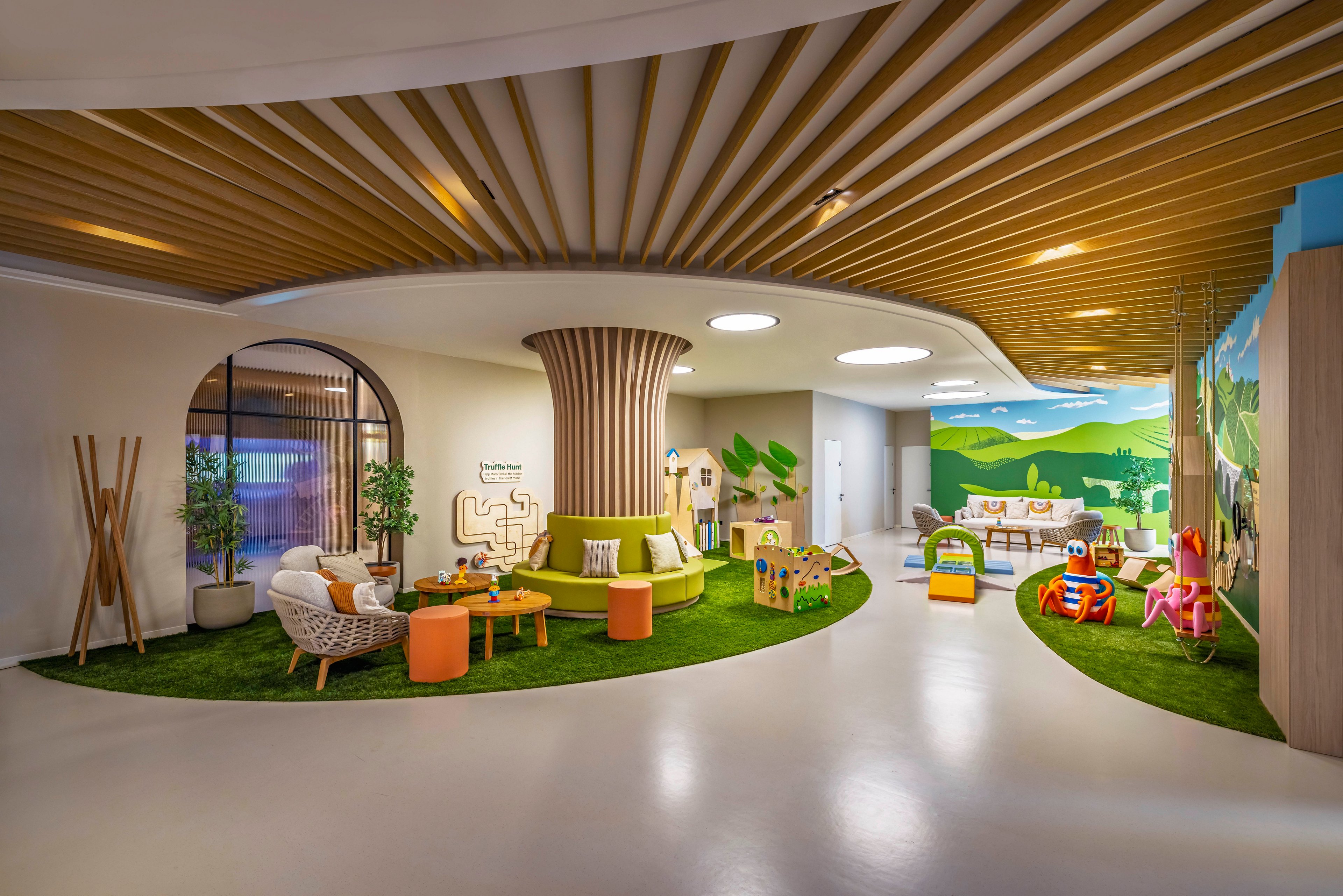 A spacious, contemporary children's playroom features playful furniture and vibrant wall murals depicting green hills and trees. The setting includes various activity zones with colorful chairs, tables, and play structures, all arranged on artificial grass mats. The ceiling design mimics wooden tree branches, enhancing the nature-inspired atmosphere. The room is well-lit with circular ceiling lights and offers a welcoming, creative environment for young children.