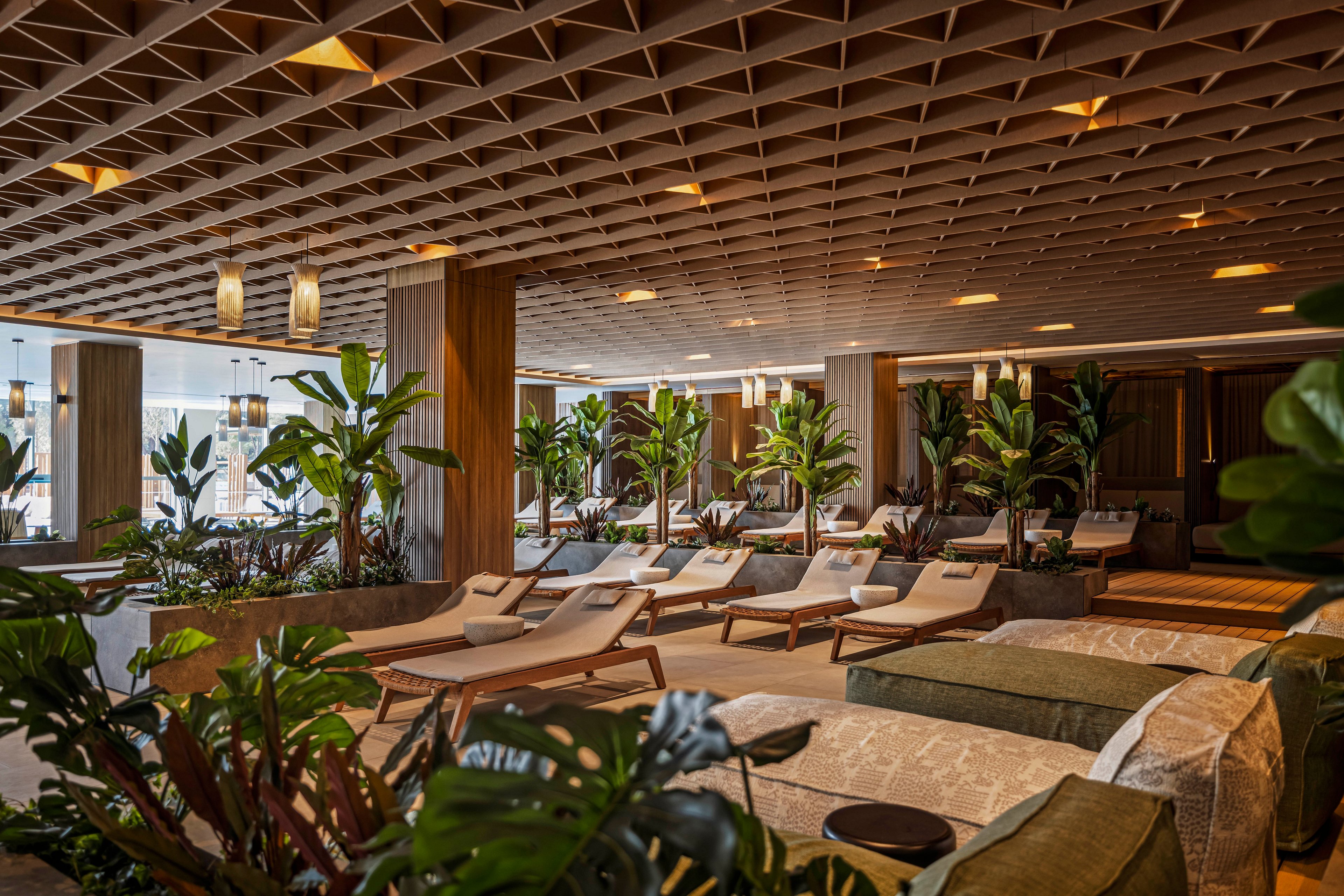 The image features a spacious indoor lounge area with multiple sunbeds arranged in rows. The setting is enhanced by abundant green plants and wooden columns, creating a relaxing and natural atmosphere. Warm lighting fixtures hang from a geometric ceiling, contributing to a cozy and inviting mood. The palette is earthy, with beige, brown, and green tones dominating the scene. No visible text or numeric values are present in the image.
