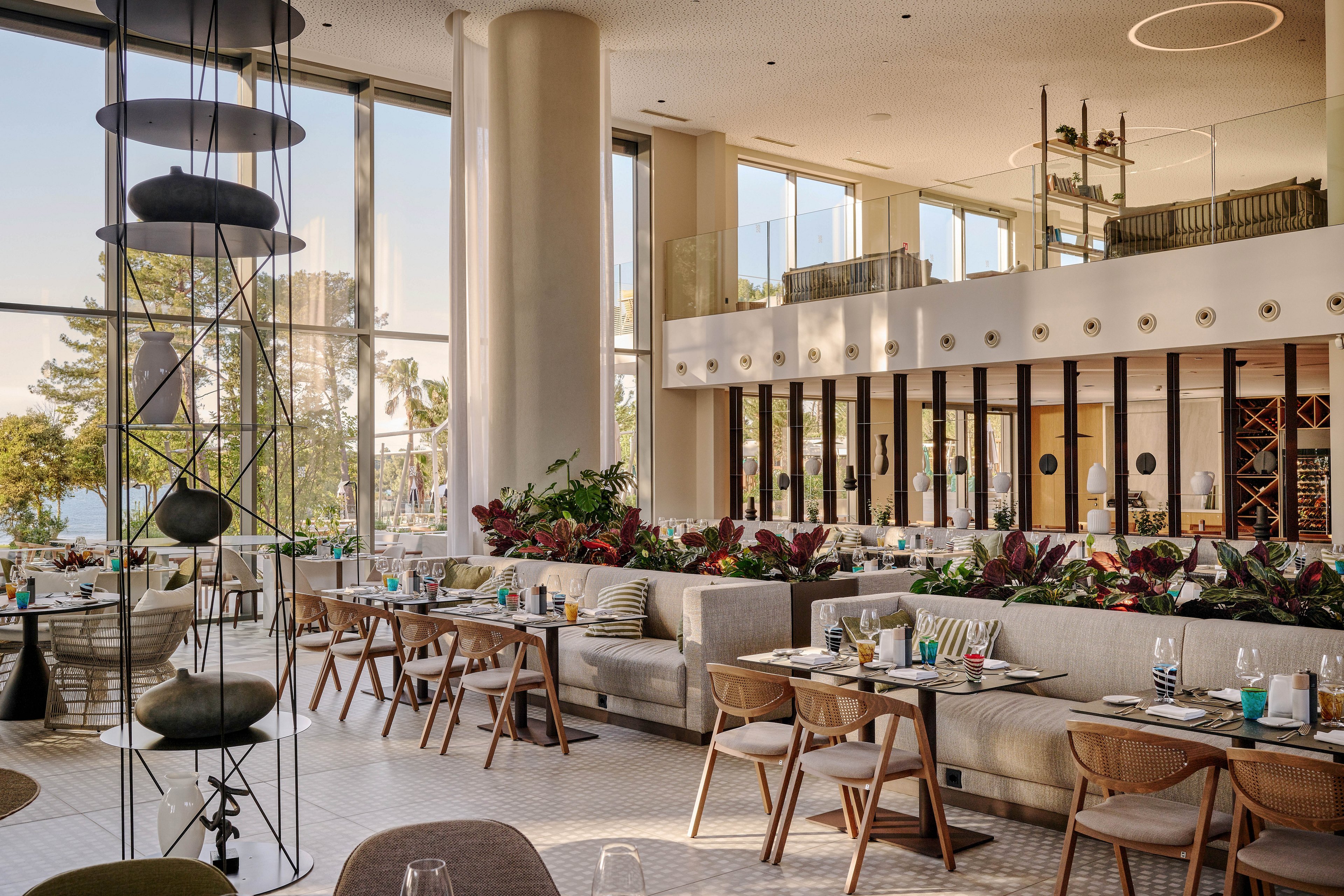 A contemporary restaurant interior features neatly arranged tables and cushioned banquettes in a bright, double-height space. Large floor-to-ceiling windows let in abundant natural light and offer views of trees outside. Green plants and red-toned foliage divide seating areas, adding a lush garden feel. Neutral tones, wooden chairs, and minimalist decor create a relaxed yet upscale dining atmosphere.