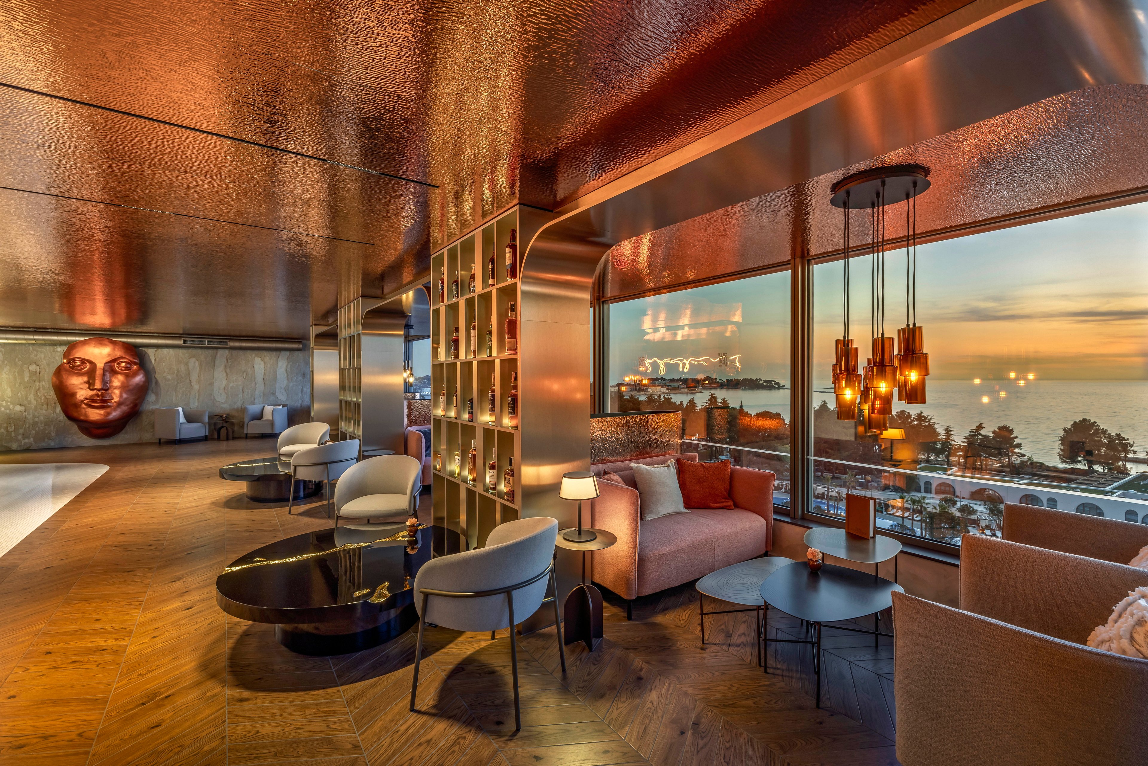Upscale lounge area features modern seating and warm ambient lighting. Large floor-to-ceiling windows reveal a panoramic ocean view at sunset. Contemporary pendant lights and reflective metallic surfaces create a sophisticated atmosphere. Shelving, side tables, and plush sofas suggest a comfortable space for relaxation or socializing.