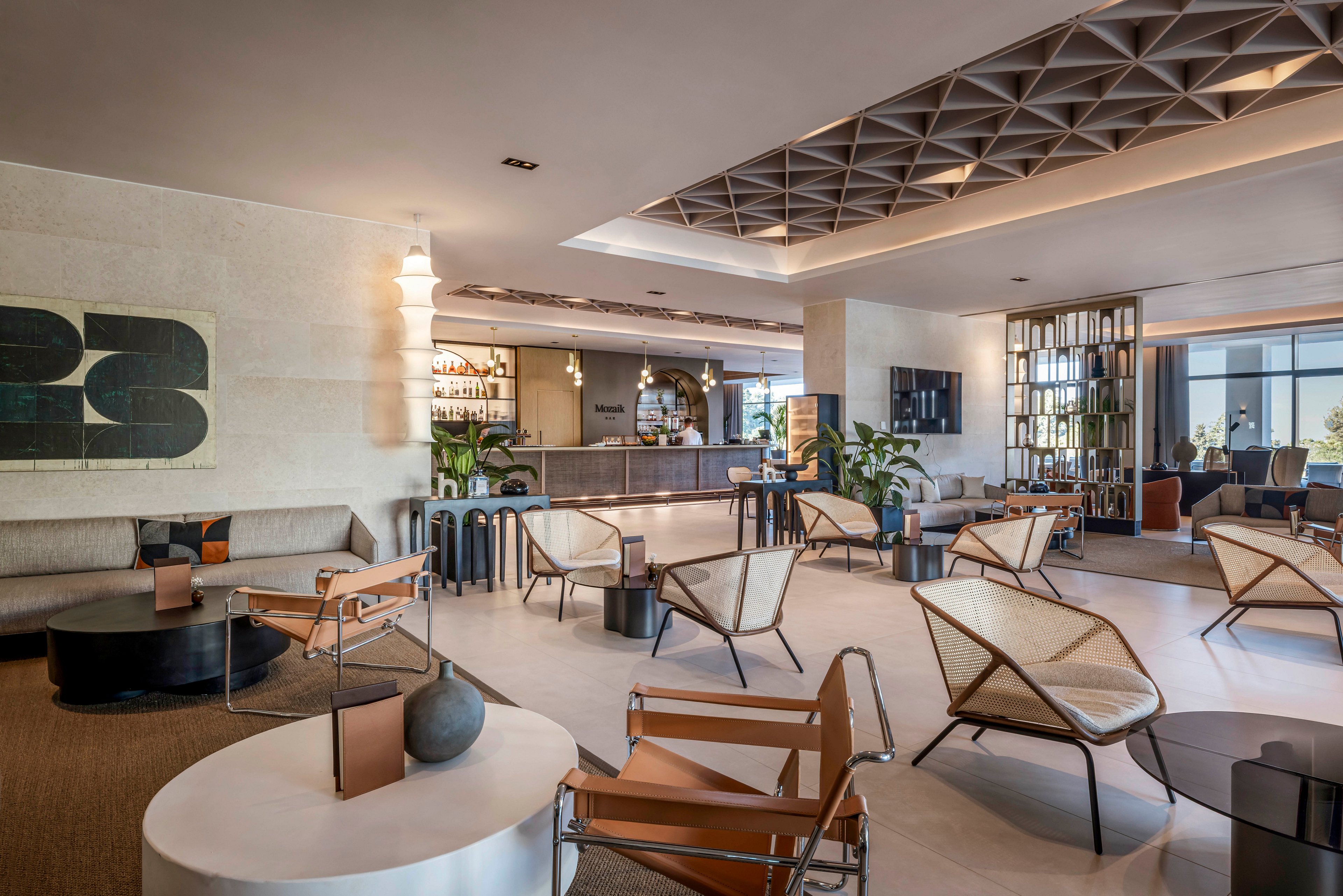 Contemporary lounge area features stylish armchairs and low tables in a spacious hotel lobby. A sleek bar with bottles and glassware is visible in the background, framed by warm lighting and architectural ceiling details. Neutral tones, clean lines, and minimalist decor create a sophisticated, welcoming atmosphere. Large windows and indoor plants add brightness and a touch of nature to the modern interior design.