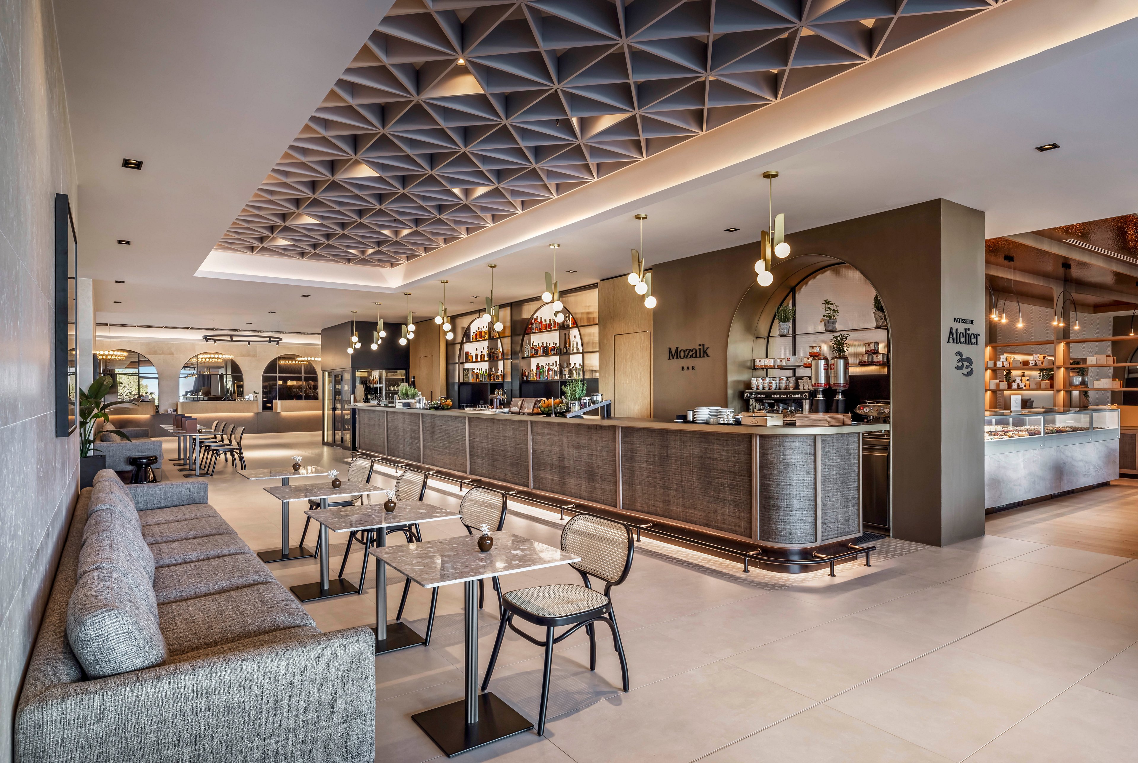 Contemporary café bar interior featuring a long counter and open shelving with bottles and glassware. The space includes cushioned bench seating and small marble-top tables with metal-framed chairs. Warm pendant lights and a geometric ceiling design create an upscale, inviting atmosphere. Neutral tones of beige, gray, and wood accents emphasize a clean, modern aesthetic. Visible wall text includes the words "Mosaic" and "Atrium" near the bar area.
