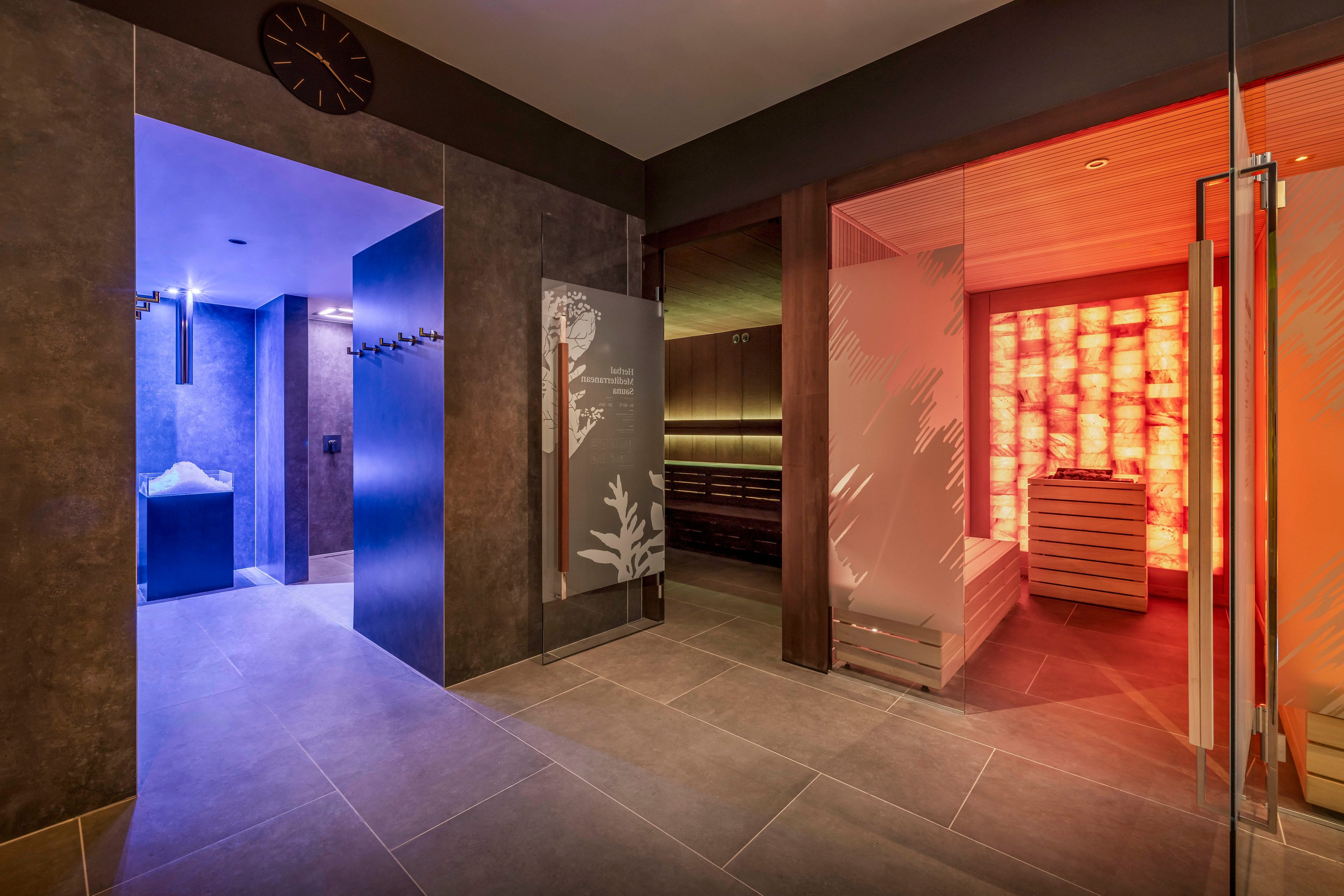 Contemporary spa interior features a corridor leading to multiple wellness rooms. On the left, a blue-lit area with a basin of ice suggests a cold therapy or ice fountain space. On the right, a warm red-orange illuminated sauna room with wooden benches creates a contrasting atmosphere. Frosted glass doors with decorative patterns and neutral floor tiles enhance the modern, relaxing design.