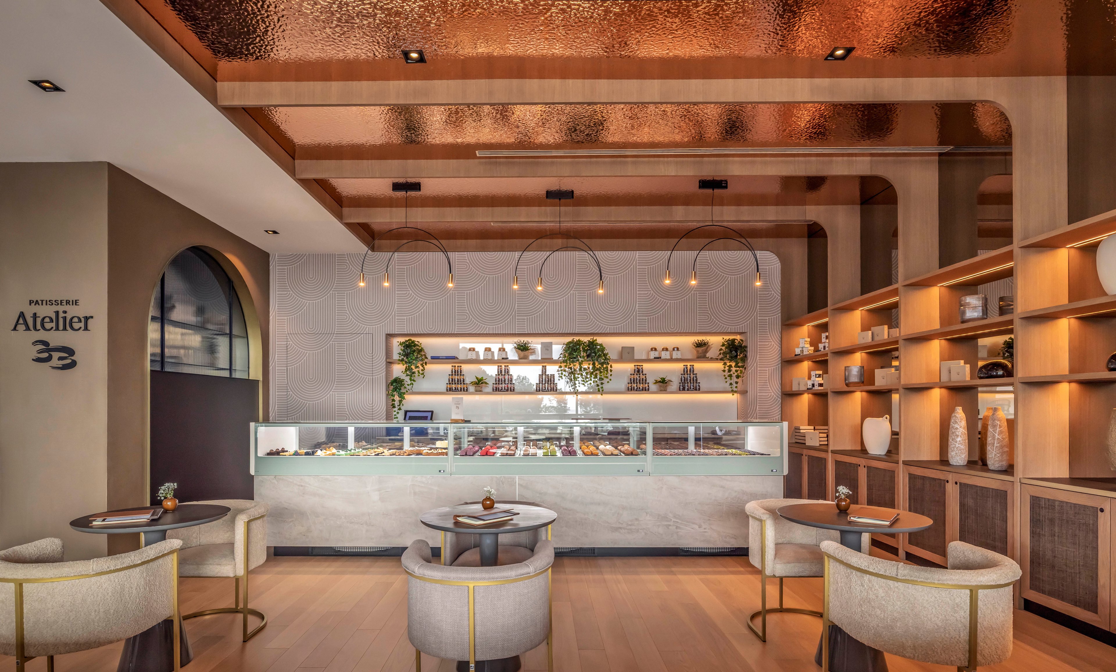 The image shows a contemporary patisserie named Atelier 33, featuring a stylish seating area with round tables and plush chairs. The setting includes a display counter filled with pastries, decorative shelving, and warm lighting. The copper ceiling and neutral palette create a sophisticated and inviting mood. Visible text includes 'Atelier 33' on the wall.