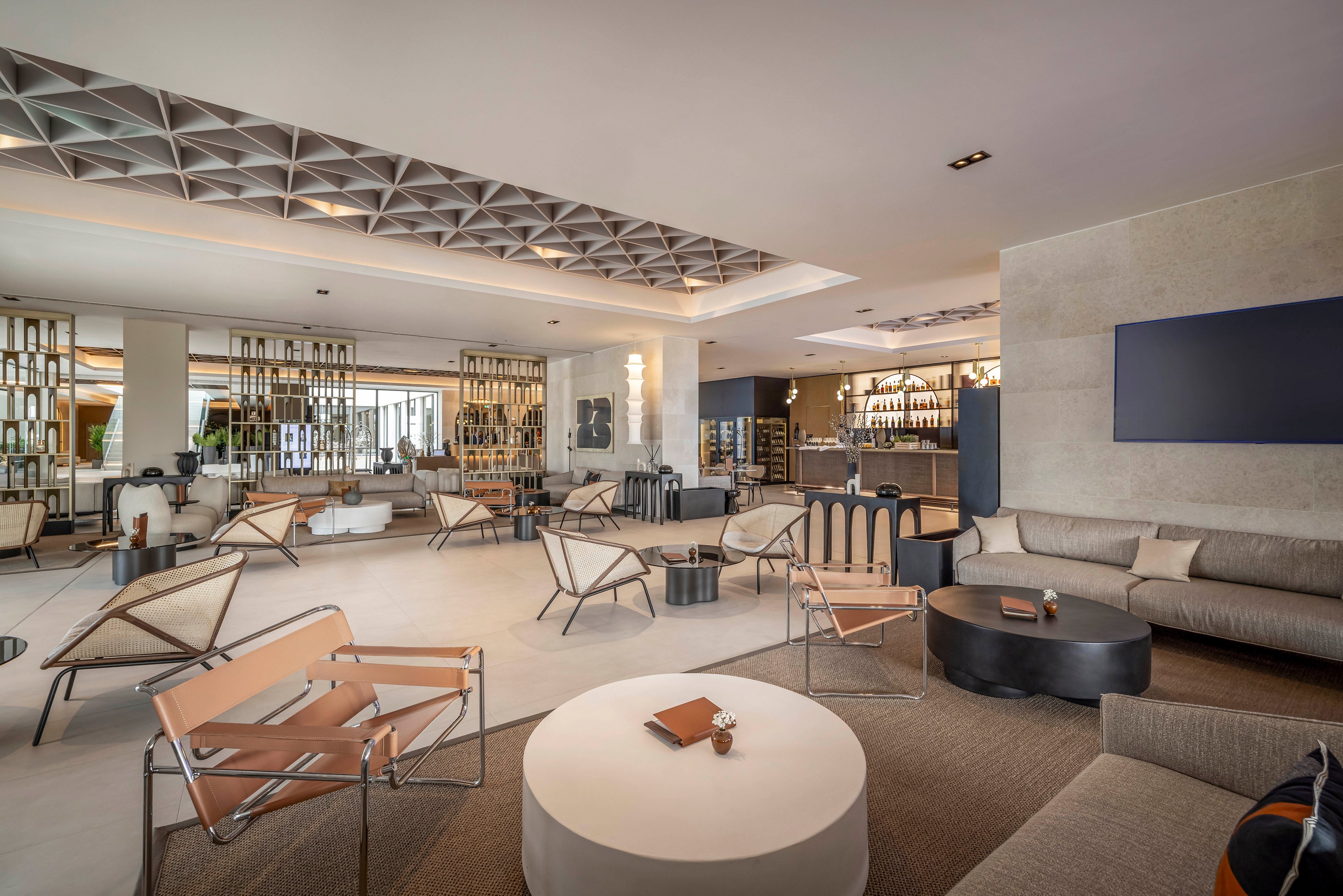 A spacious hotel lounge features contemporary furniture, including minimalist chairs and sofas arranged around low tables. The setting is bright and airy, with neutral tones and geometric ceiling details enhancing the modern aesthetic. Shelving with decorative items and a well-lit bar area are visible in the background. The overall mood is calm and inviting, ideal for relaxation or informal meetings.