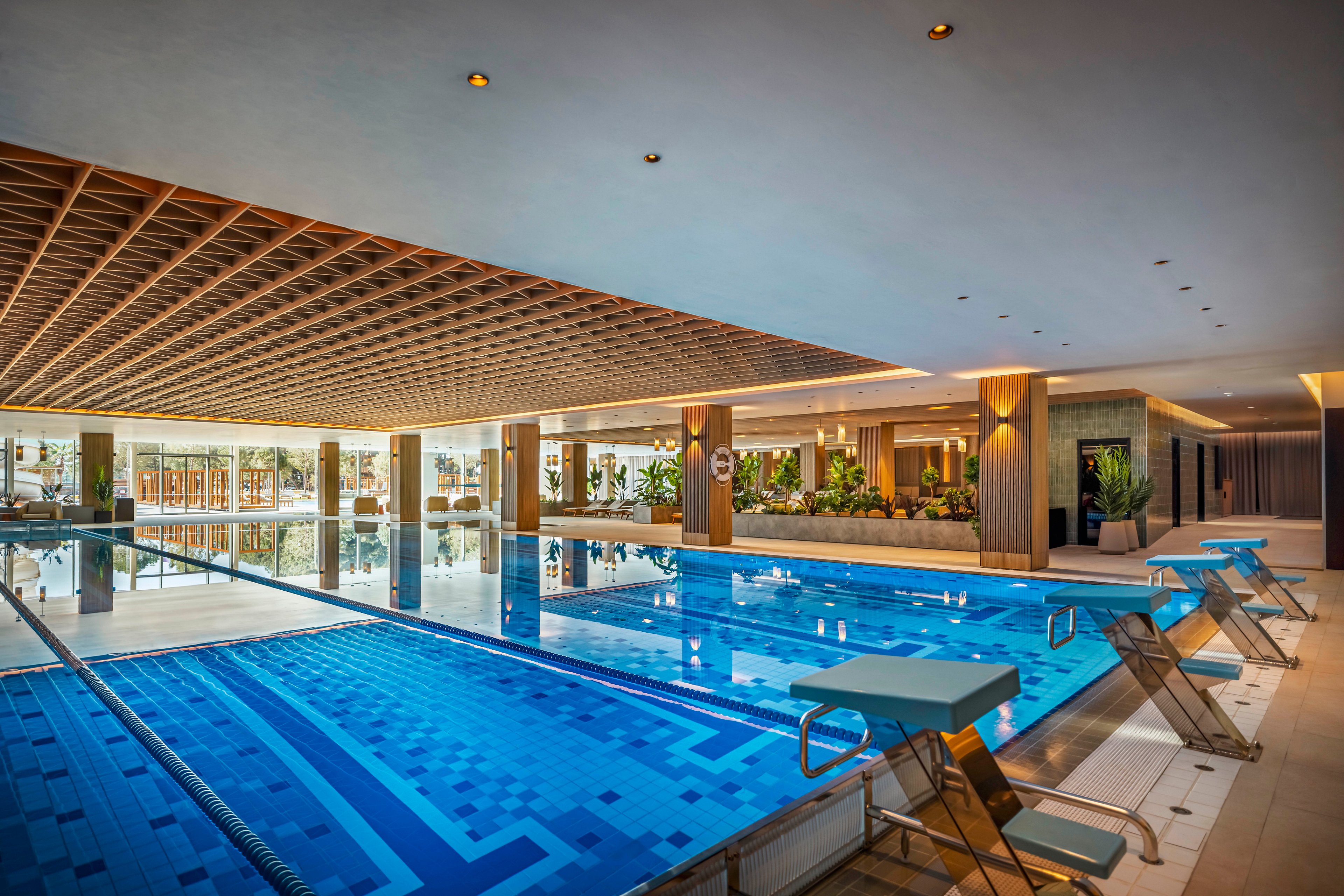 A spacious indoor swimming pool is featured in a contemporary leisure facility. The pool includes multiple lanes and several starting blocks, with clear blue water and geometric tile patterns. The setting is well-lit with natural light from large windows, and the area is accented by wooden ceiling panels and lush green plants. No visible people or text are present in the image.