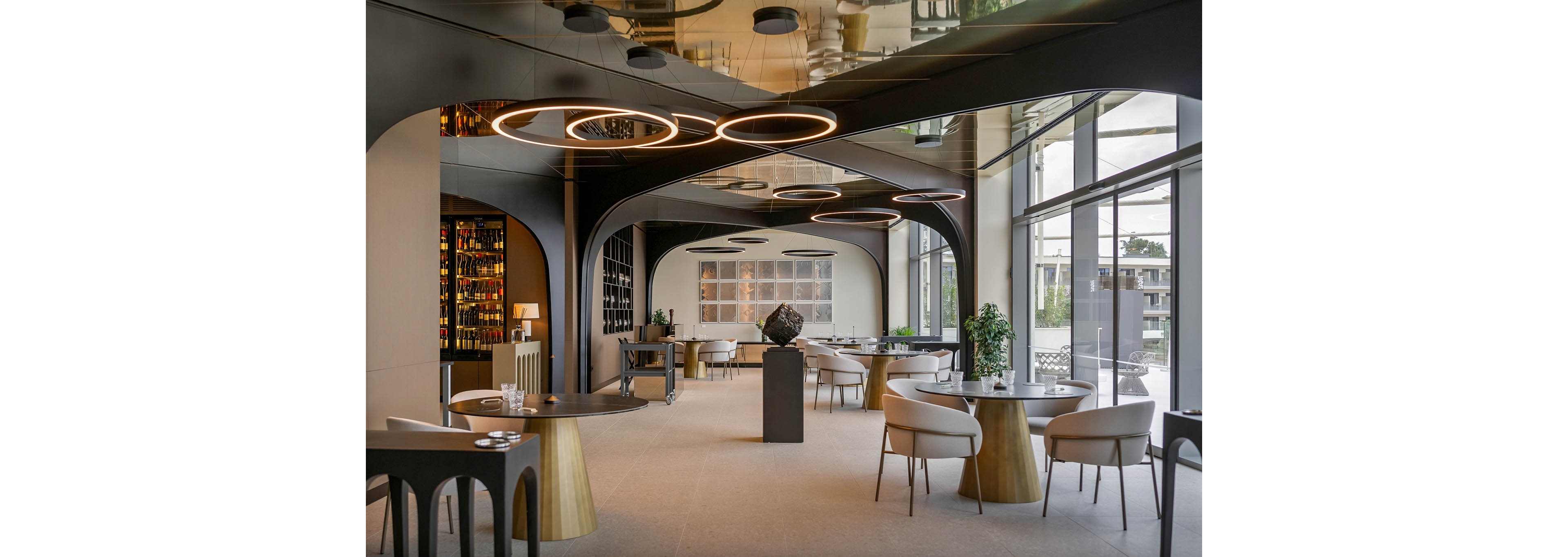 A contemporary restaurant dining area features sleek circular pendant lights and minimalist furniture. The space is filled with natural light from large floor-to-ceiling windows, offering a view of the outdoors. Elegant tables and chairs are arranged throughout, with decorative plants and a geometric sculpture adding visual interest. The overall mood is sophisticated and inviting, with a neutral color palette and modern design elements.