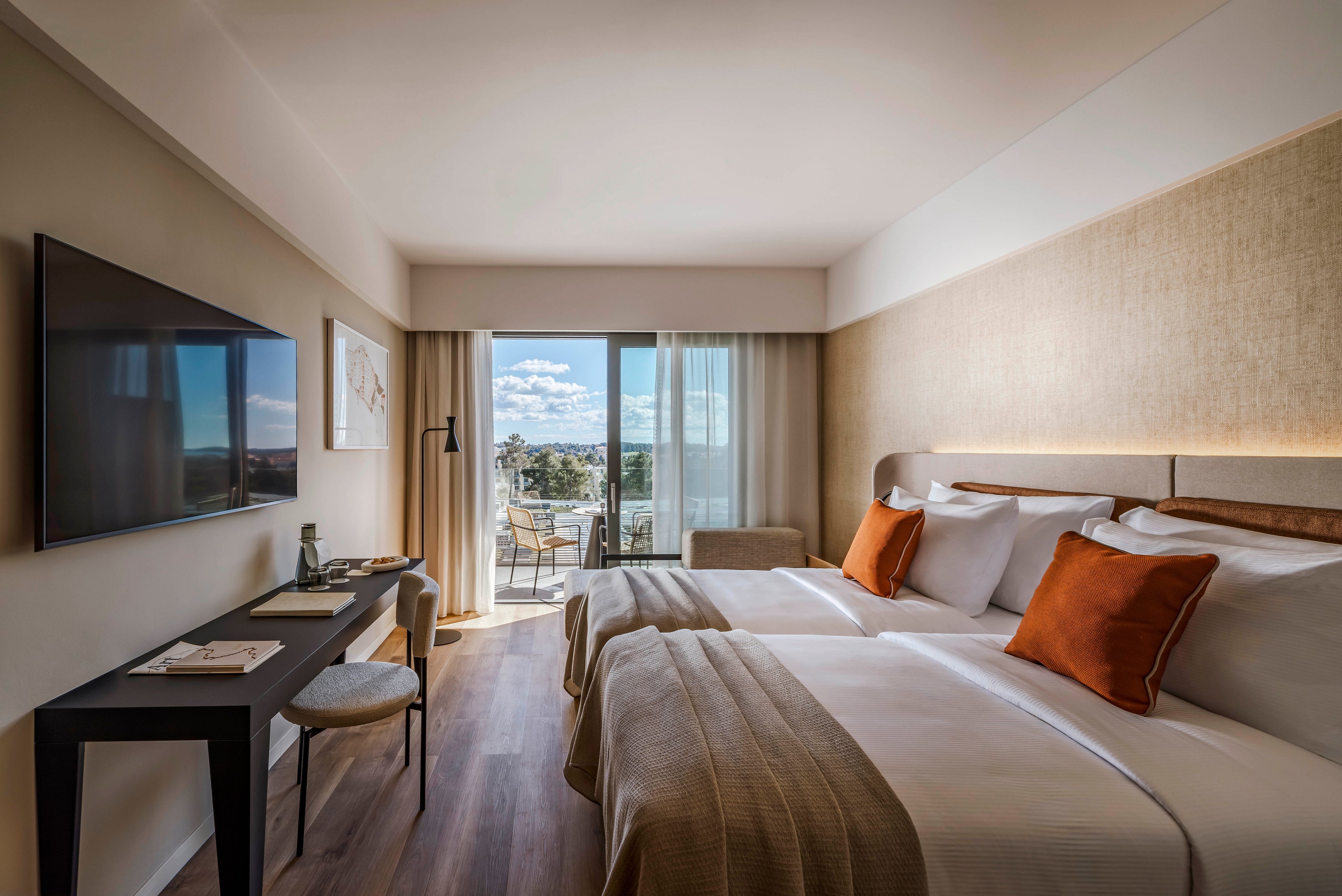 A contemporary hotel room features two neatly made beds with white linens and orange accent pillows. The space includes a wall-mounted flat-screen TV, a work desk with a chair, and a large window leading to a balcony overlooking a cityscape. Natural light fills the room, highlighting the neutral color palette and minimalist decor. The overall atmosphere is calm and inviting, ideal for relaxation or business travel.